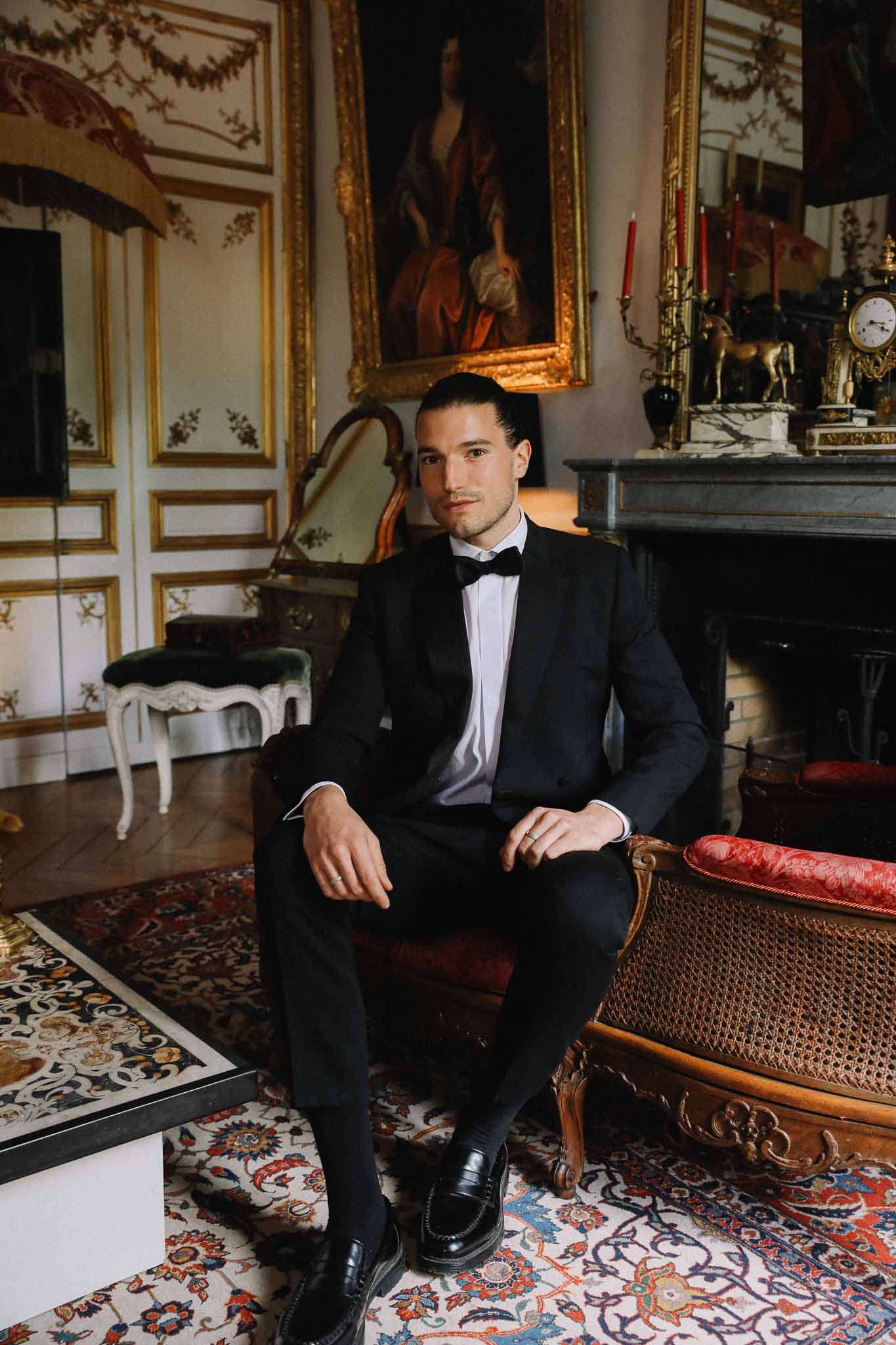 Groom in black tuxedo seated for formal portrait in ornate classical interior with gilt-framed artwork