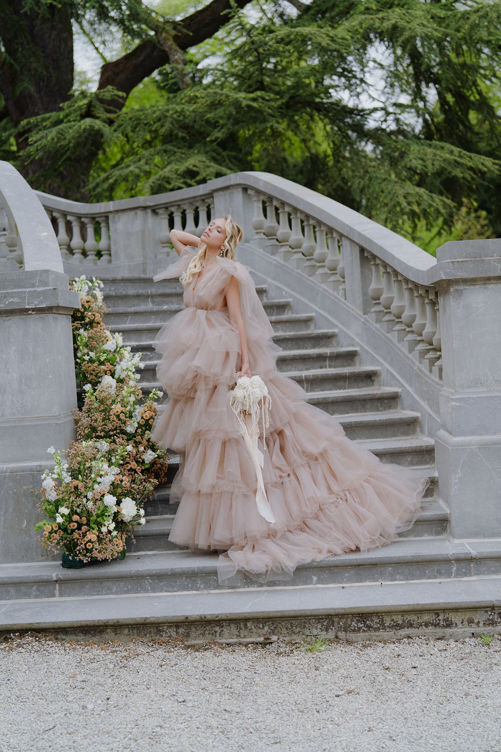 Bride in blush tiered tulle gown on chateau staircase with pearl-strand bouquet and amber floral arrangement