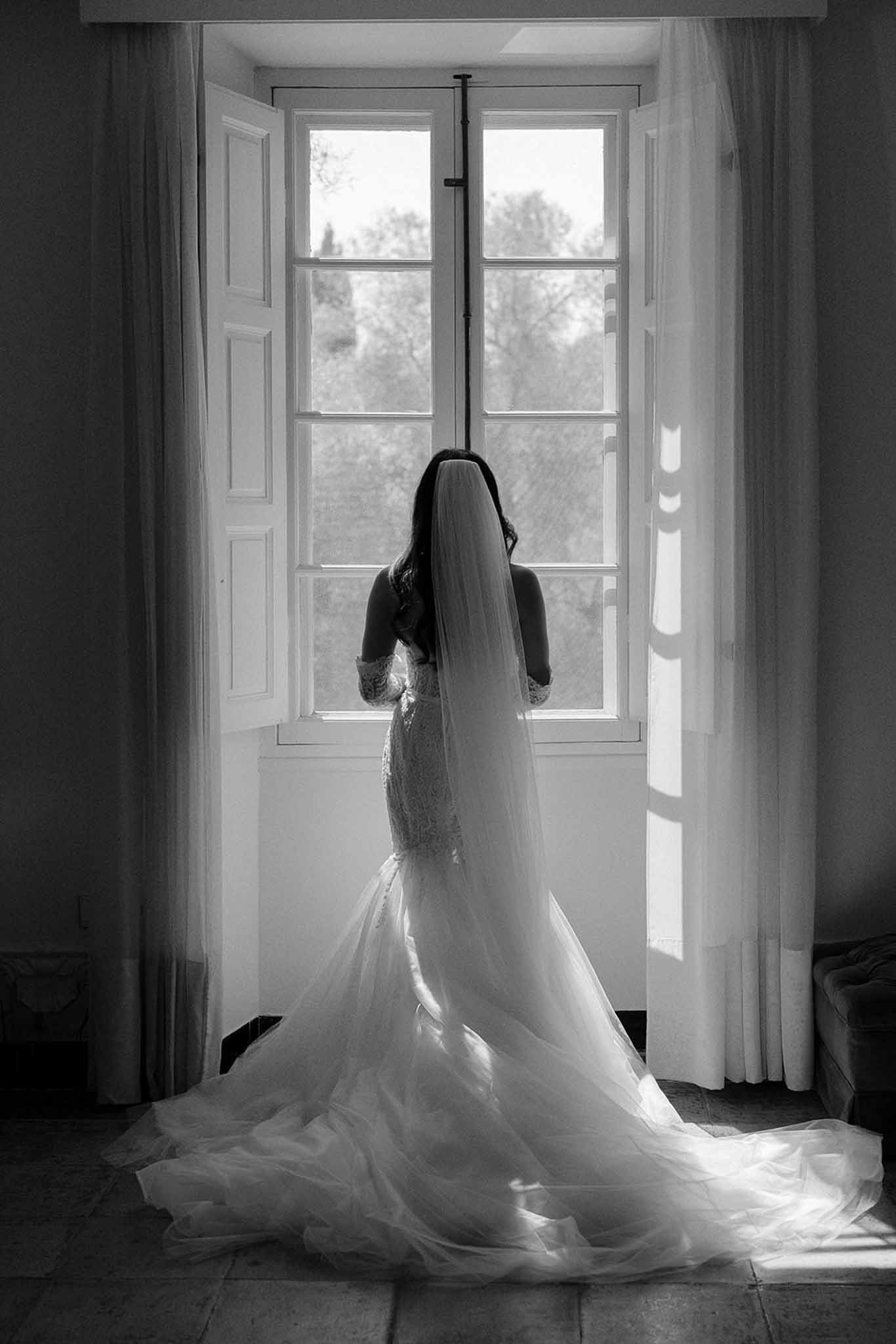 Black and white bridal portrait from behind, bride in lace mermaid gown with cathedral veil silhouetted against French win...