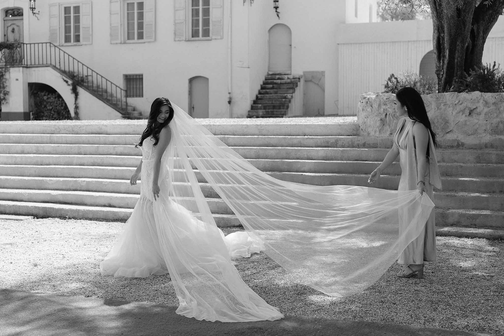 Black and white bridesmaid holding cathedral veil aloft as bride smiles on chateau gravel courtyard