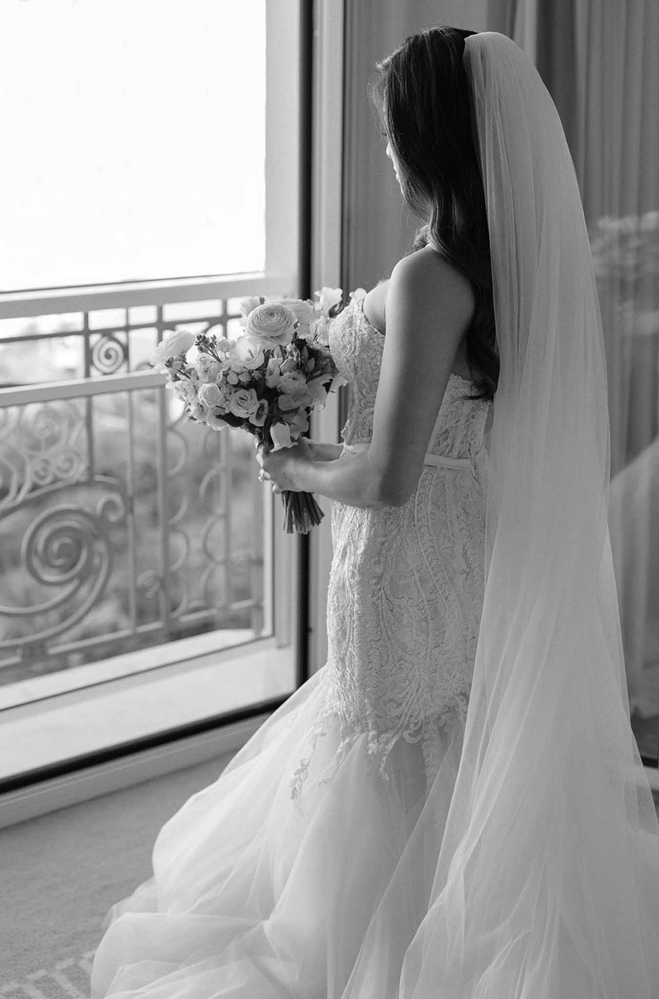 Black and white portrait of bride from behind in strapless lace mermaid gown with cathedral veil holding a lush bouquet by...