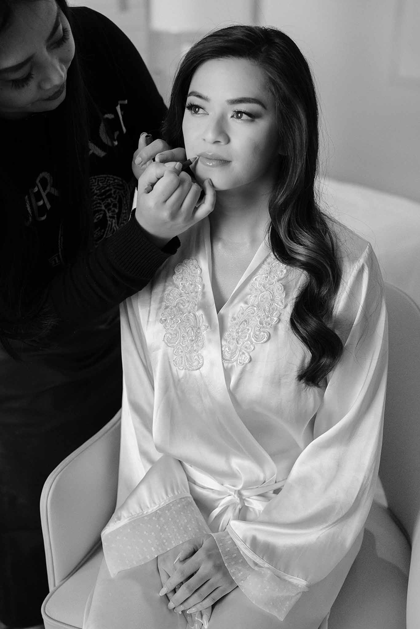 Black and white photo of a makeup artist applying lipstick to a bride wearing a lace-trimmed satin robe