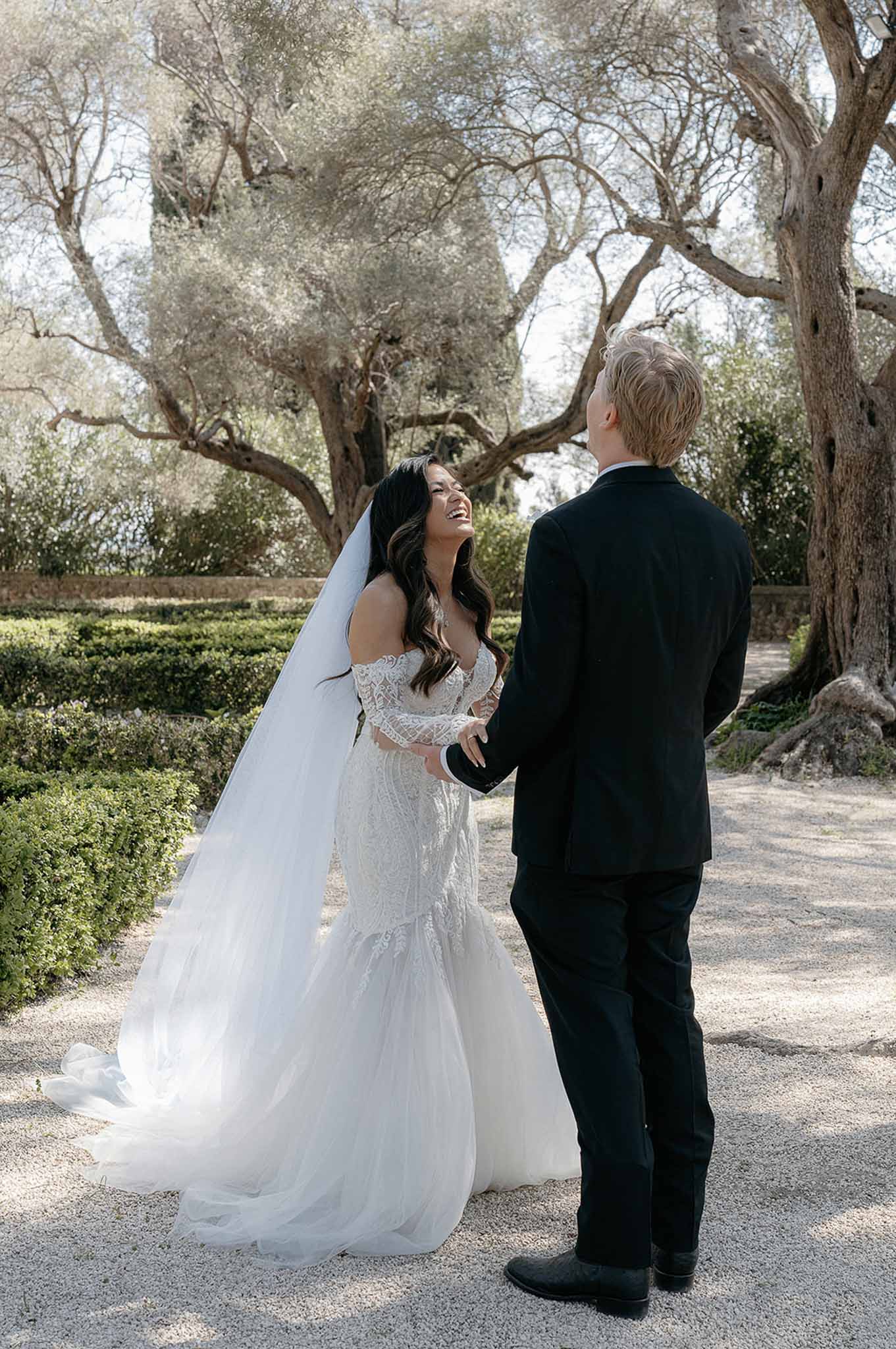 Bride in off-the-shoulder lace mermaid gown and cathedral veil laughing with groom during first look in formal garden