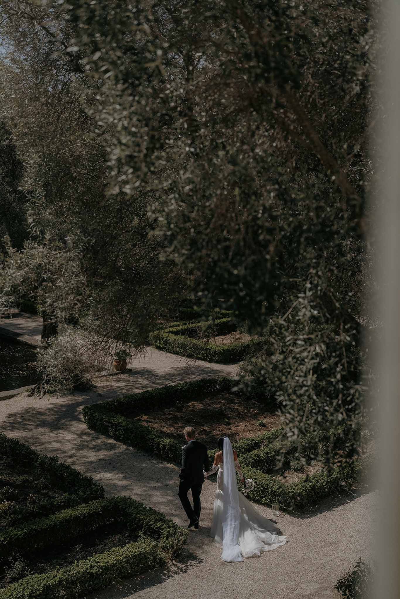 Elevated view of couple walking through box parterre garden with cathedral veil trailing on gravel path