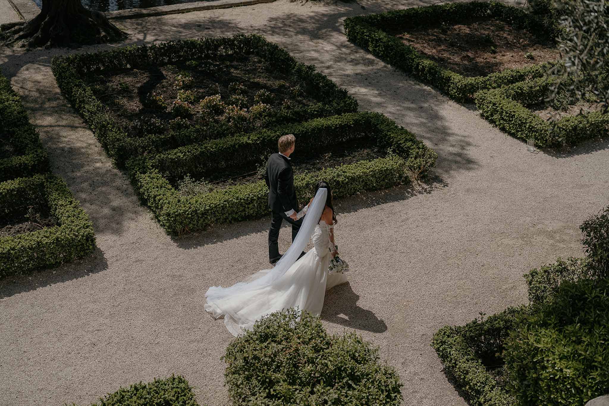 Overhead shot of bride and groom walking through formal boxwood parterre garden with cathedral train spread on gravel