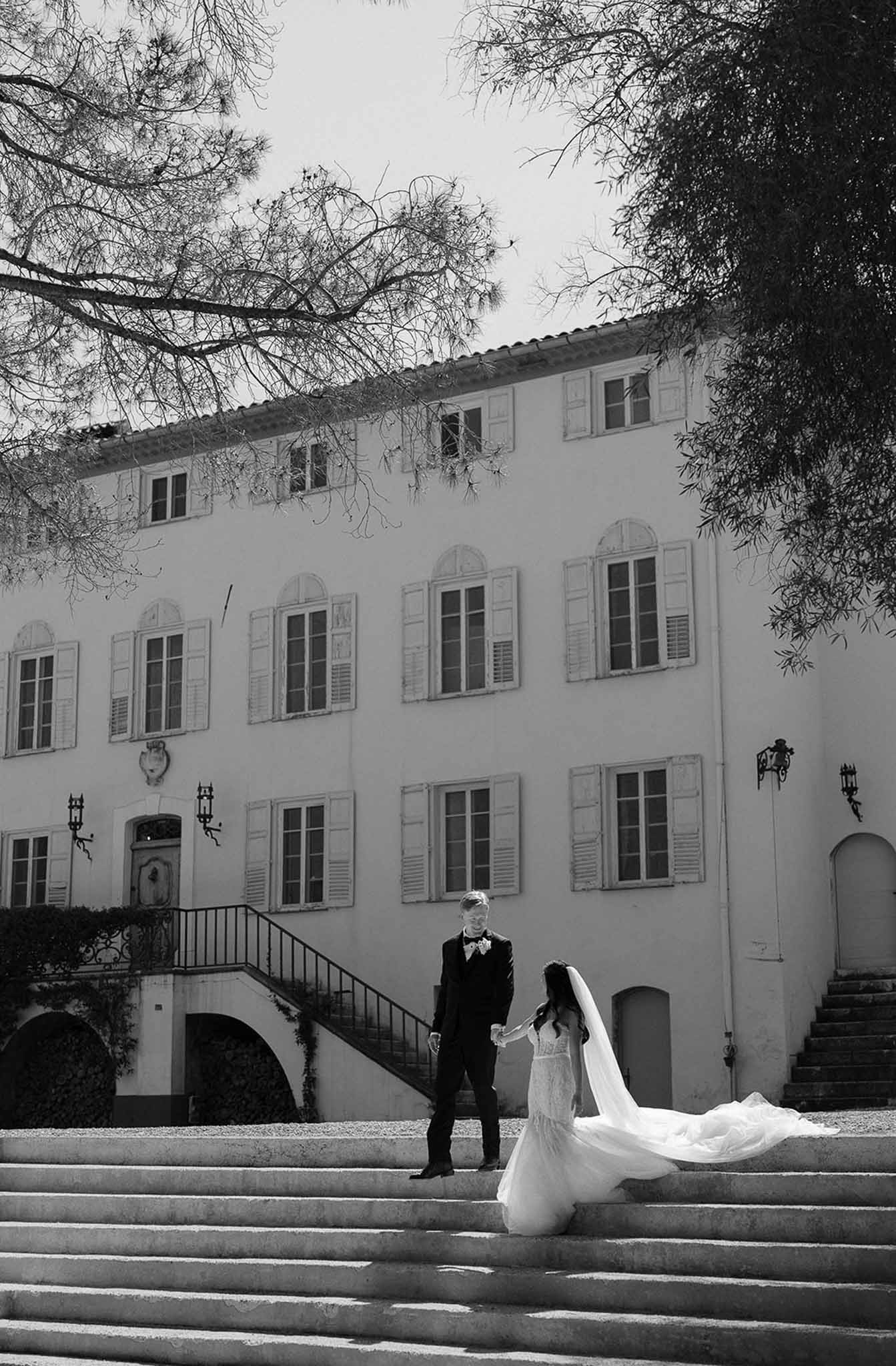 Bride with cathedral veil fanned across steps and groom hold hands before Provencal bastide in B&W