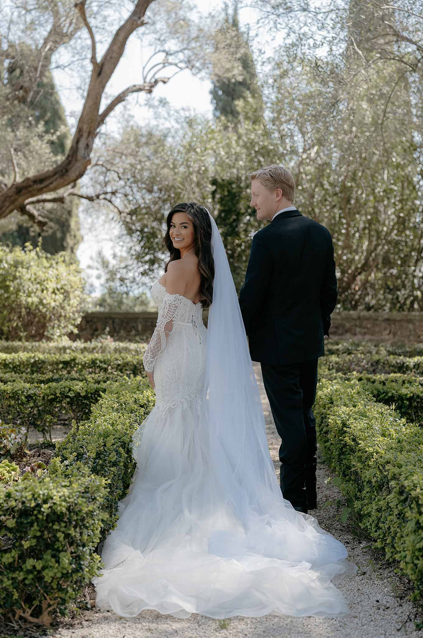 Bride in lace gown with cathedral veil and groom walking through formal French parterre garden, shot from behind