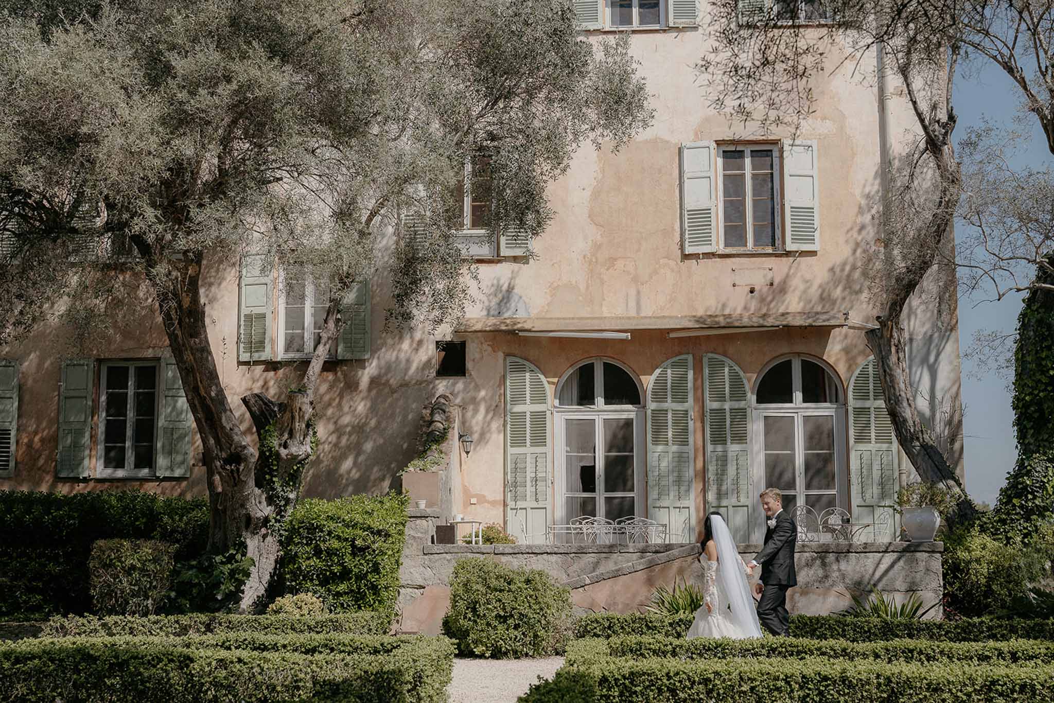 Couple walking before terracotta bastide with sage shutters bride in fitted gown with cathedral veil on box garden