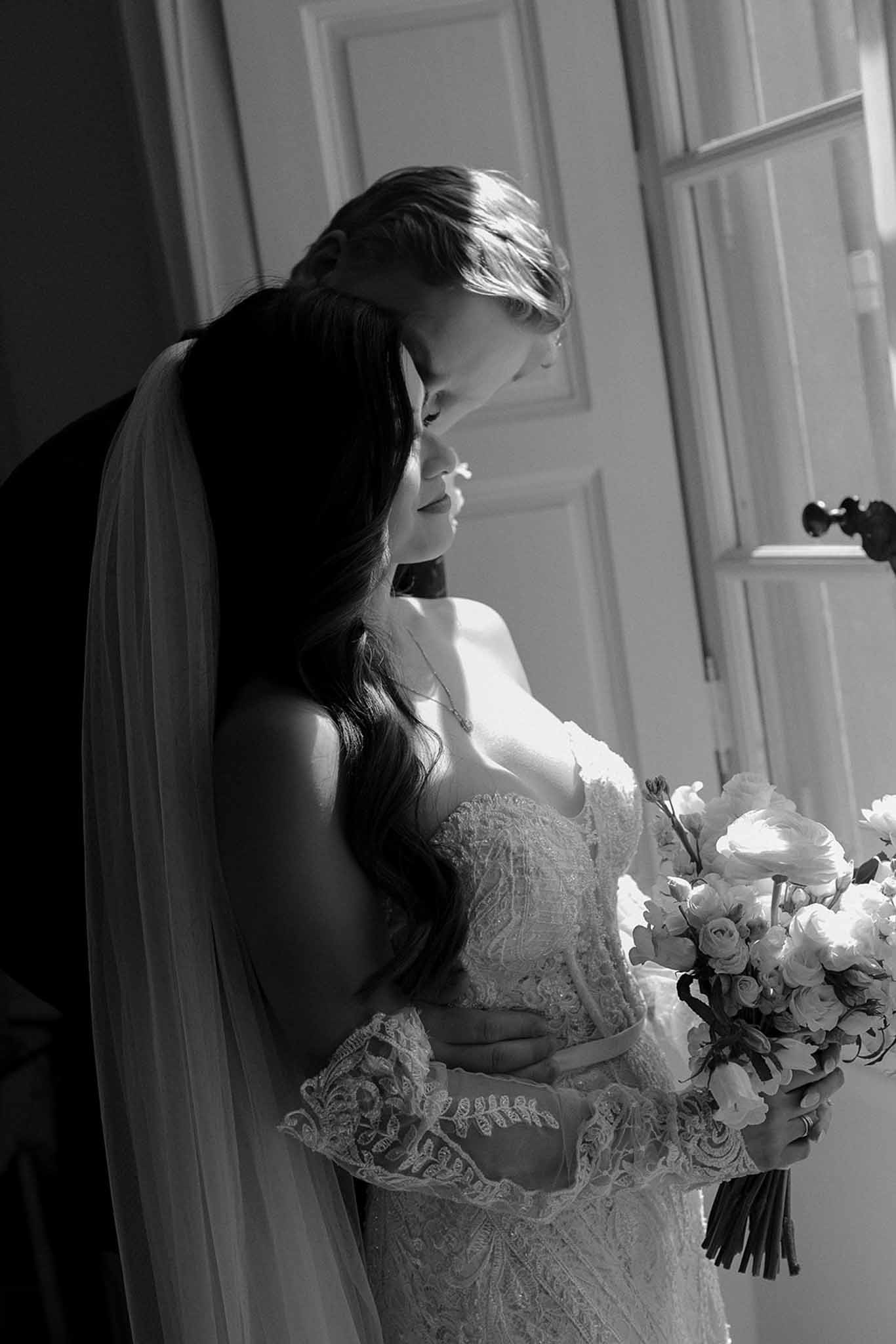 Black-and-white portrait of groom kissing bride on the head as she holds a bouquet inside a chateau doorway