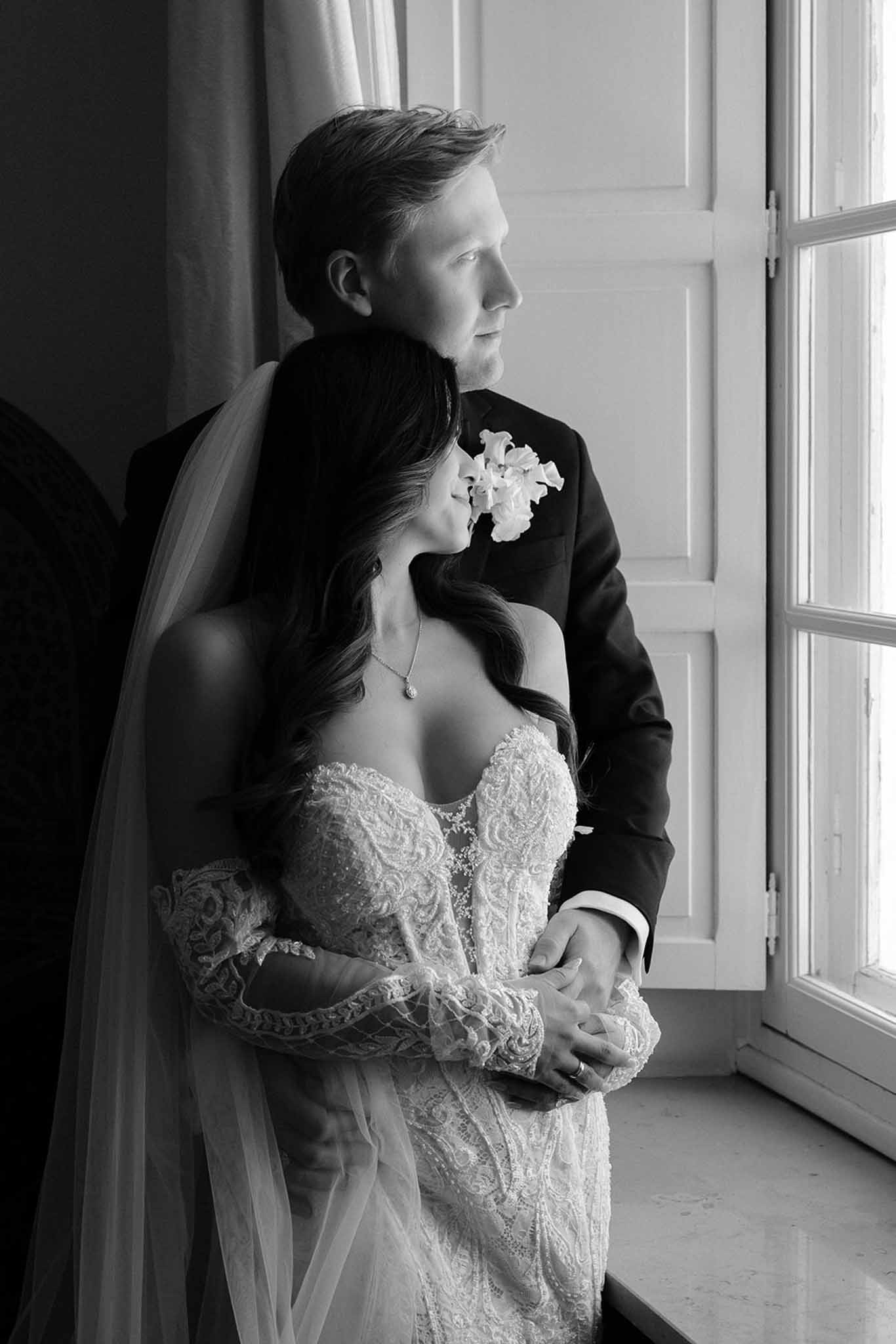 Black and white couple by French window with embroidered lace gown and orchid boutonniere
