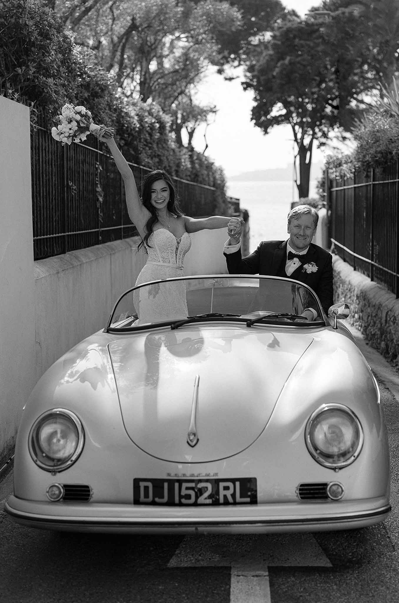 Black and white of couple celebrating in vintage Porsche Speedster, bride raising bouquet, groom with glass