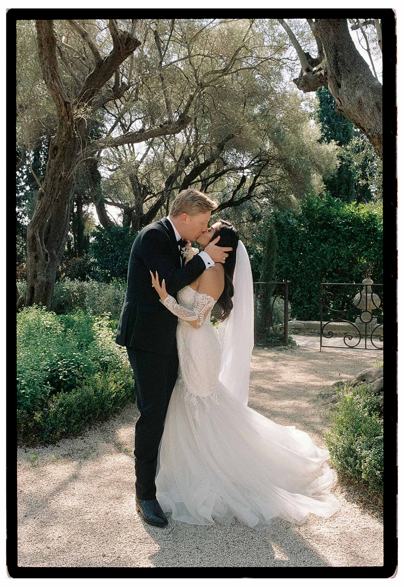 Couple kissing on garden path with ornamental gate, bride in ivory lace gown with cathedral veil catching breeze