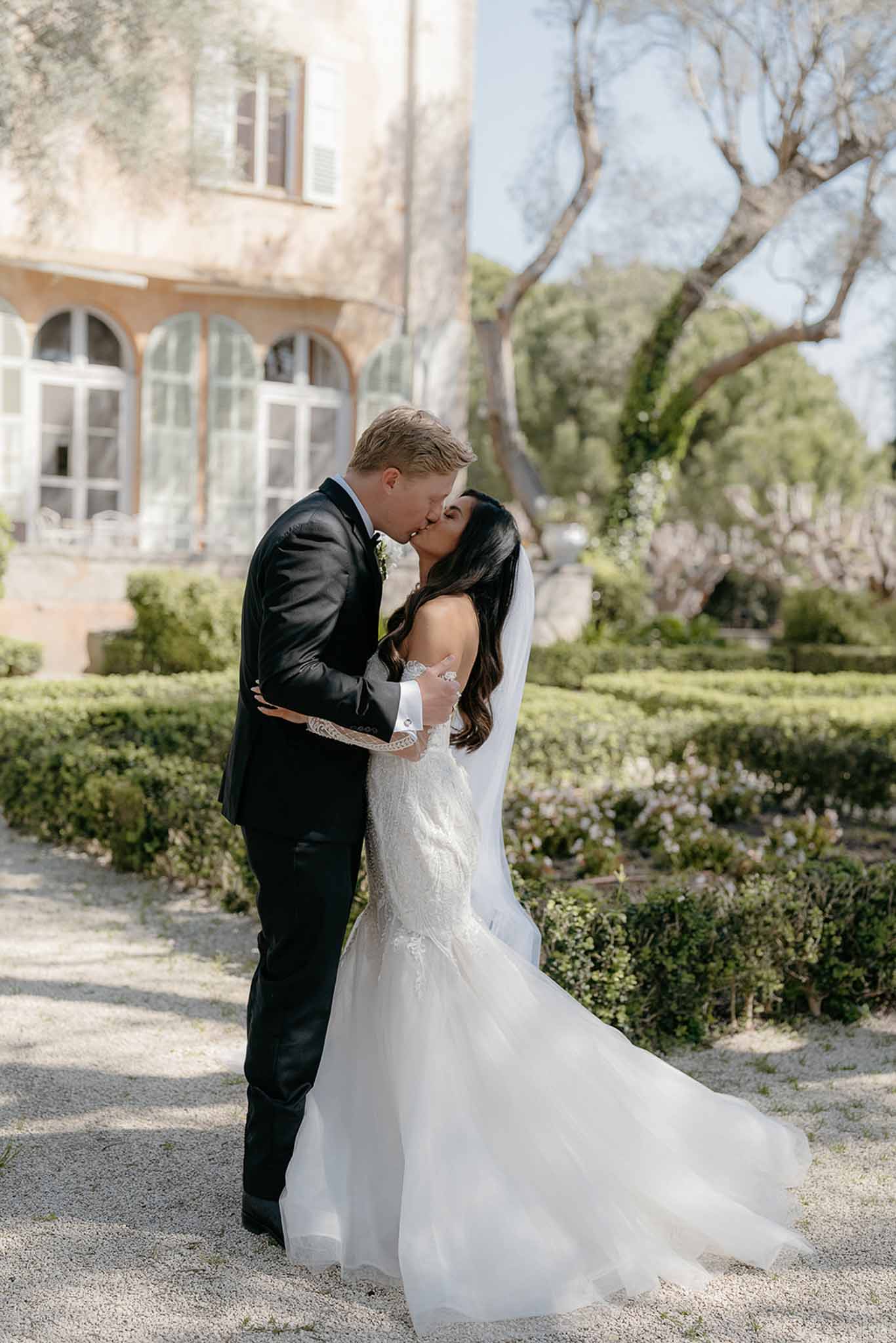 Couple kissing on gravel path in formal parterre garden bride in lace mermaid gown with cathedral veil ochre bastide behind