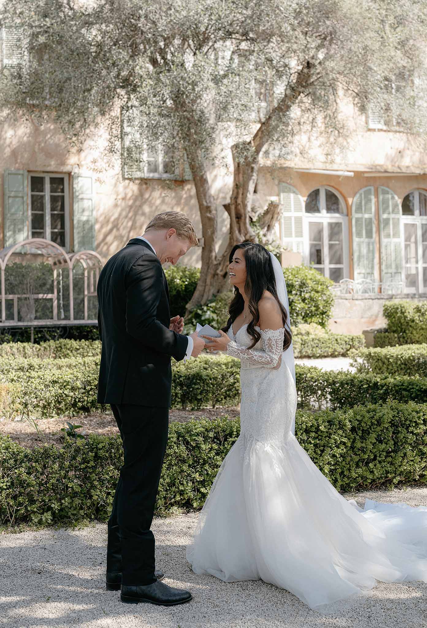 Bride in off-shoulder lace sleeve gown and groom in black suit sharing first look in chateau garden courtyard