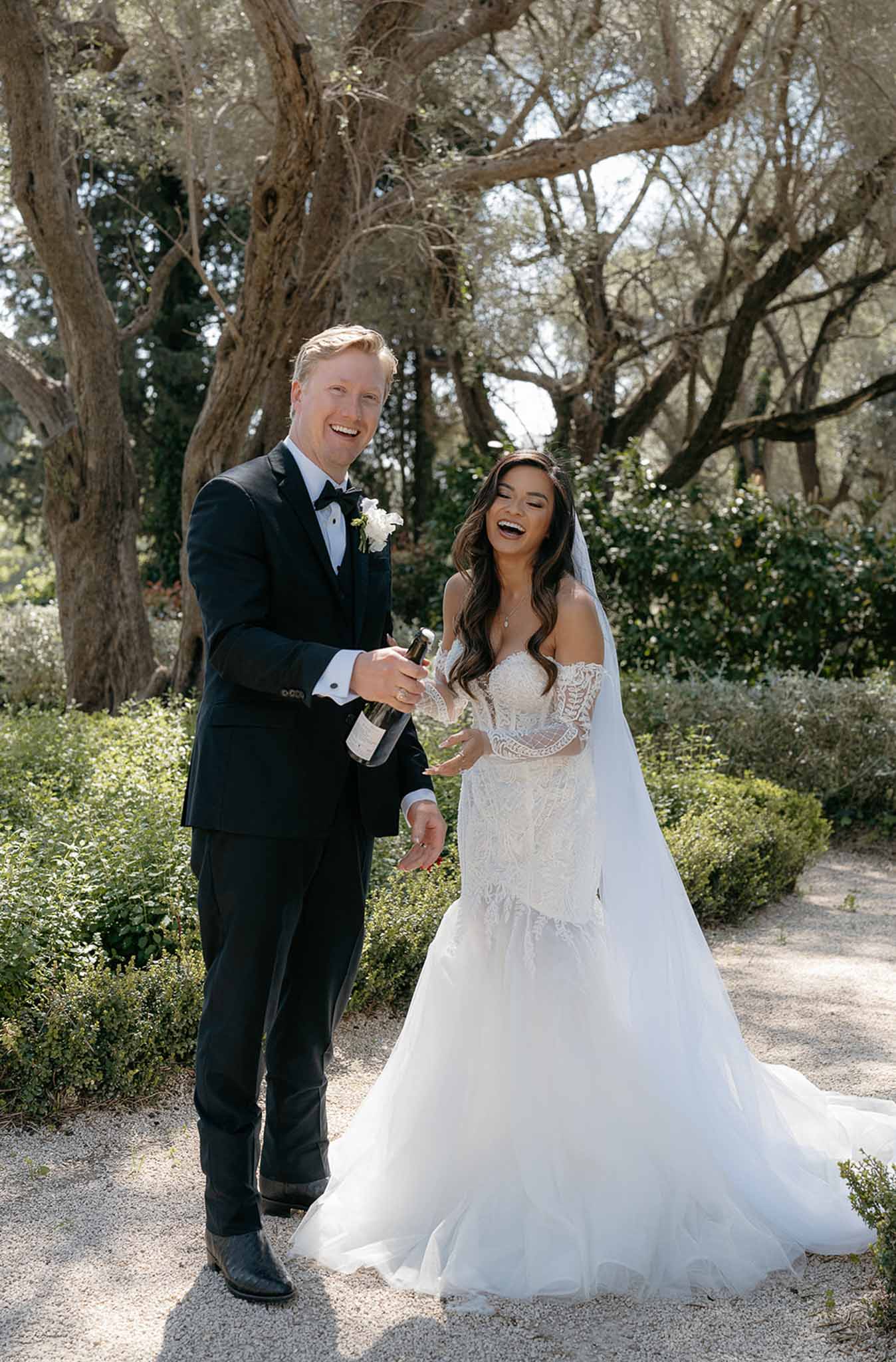 Bride and groom laughing while opening champagne bottle on gravel garden path with formal hedgerows