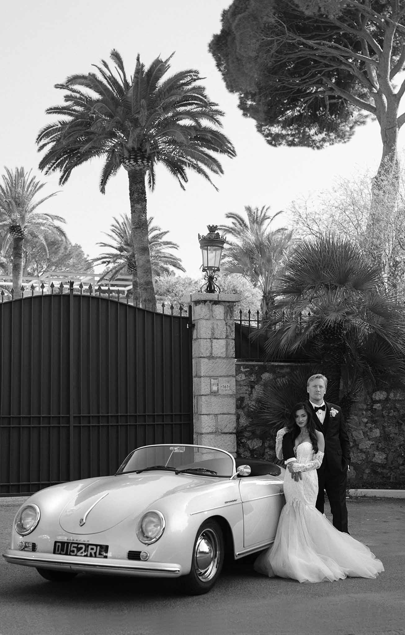 Black and white couple posing beside vintage Porsche 356 Speedster at ornate stone gate with iron lantern