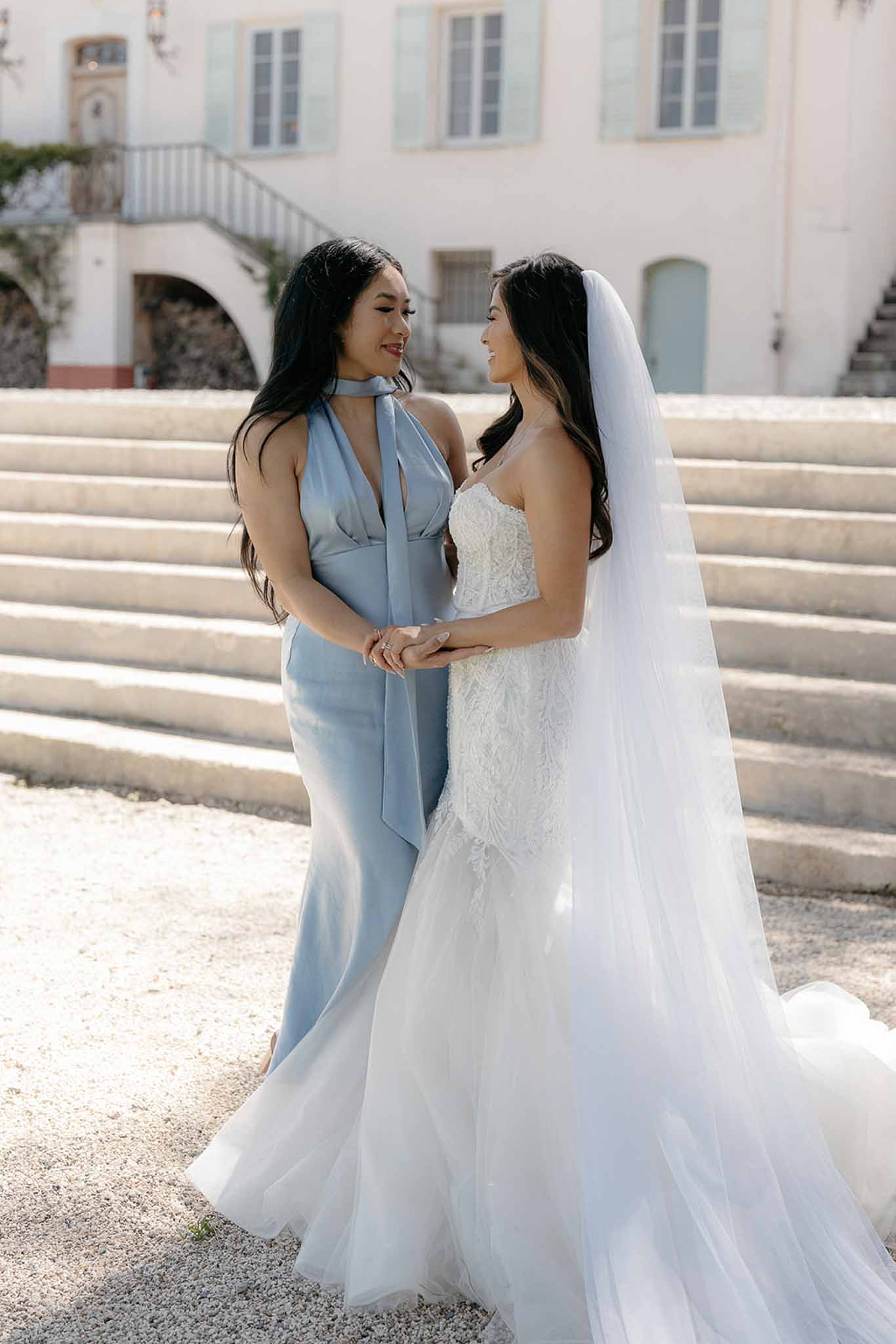 Bride in strapless lace gown and bridesmaid in dusty blue satin dress holding hands before pink manor