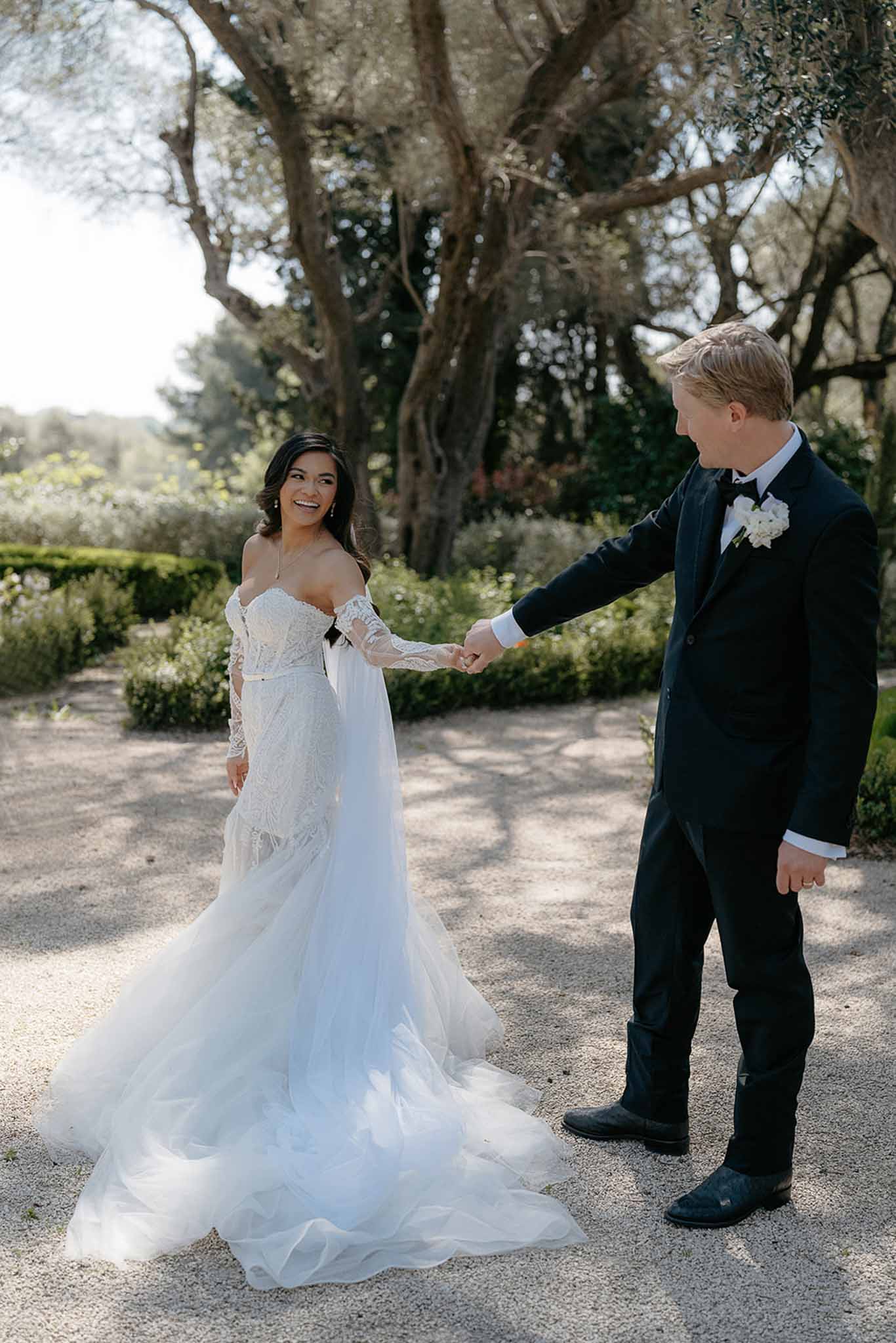 Bride in off-shoulder lace mermaid gown holding hands with groom in black tuxedo on garden path