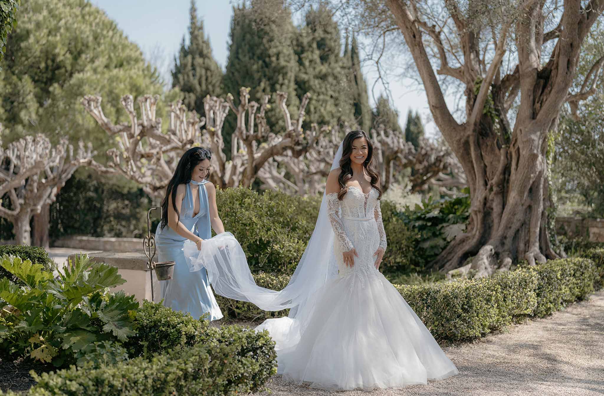 Bride in off-shoulder lace mermaid gown with cathedral train as bridesmaid in blue adjusts veil in garden