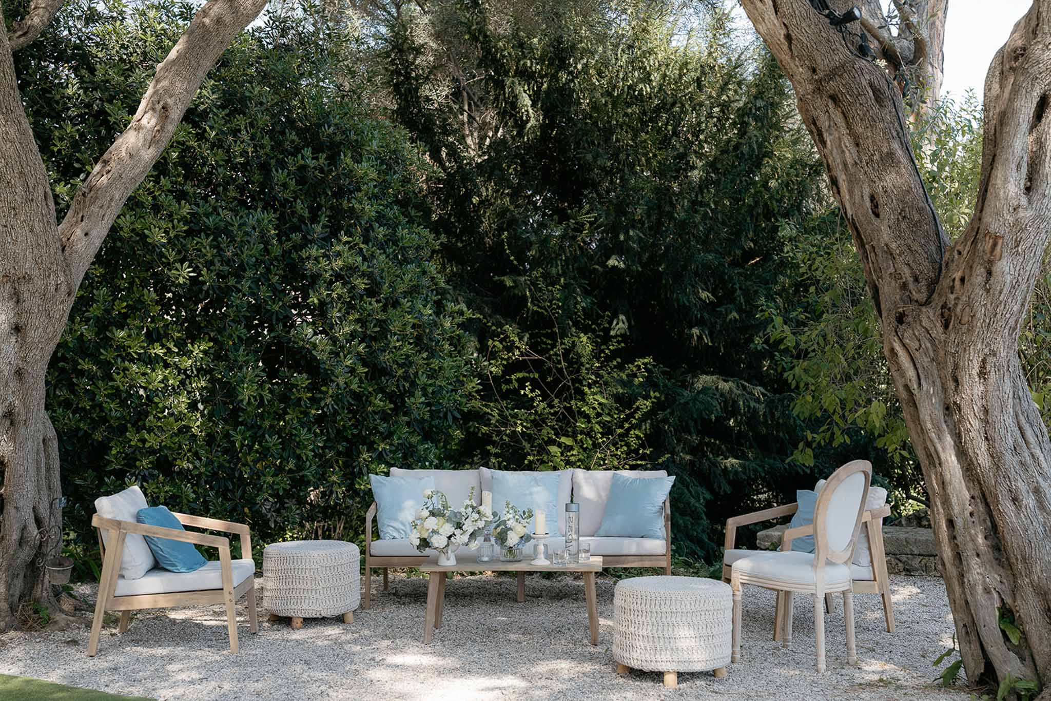 Outdoor lounge area with cream sofas and blue pillows between olive trees, low table with white floral arrangements