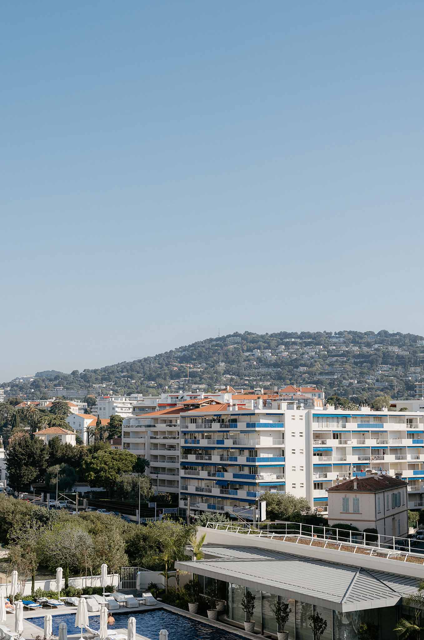 Elevated view of hotel pool terrace with white loungers and glass pavilion overlooking coastal town rooftops