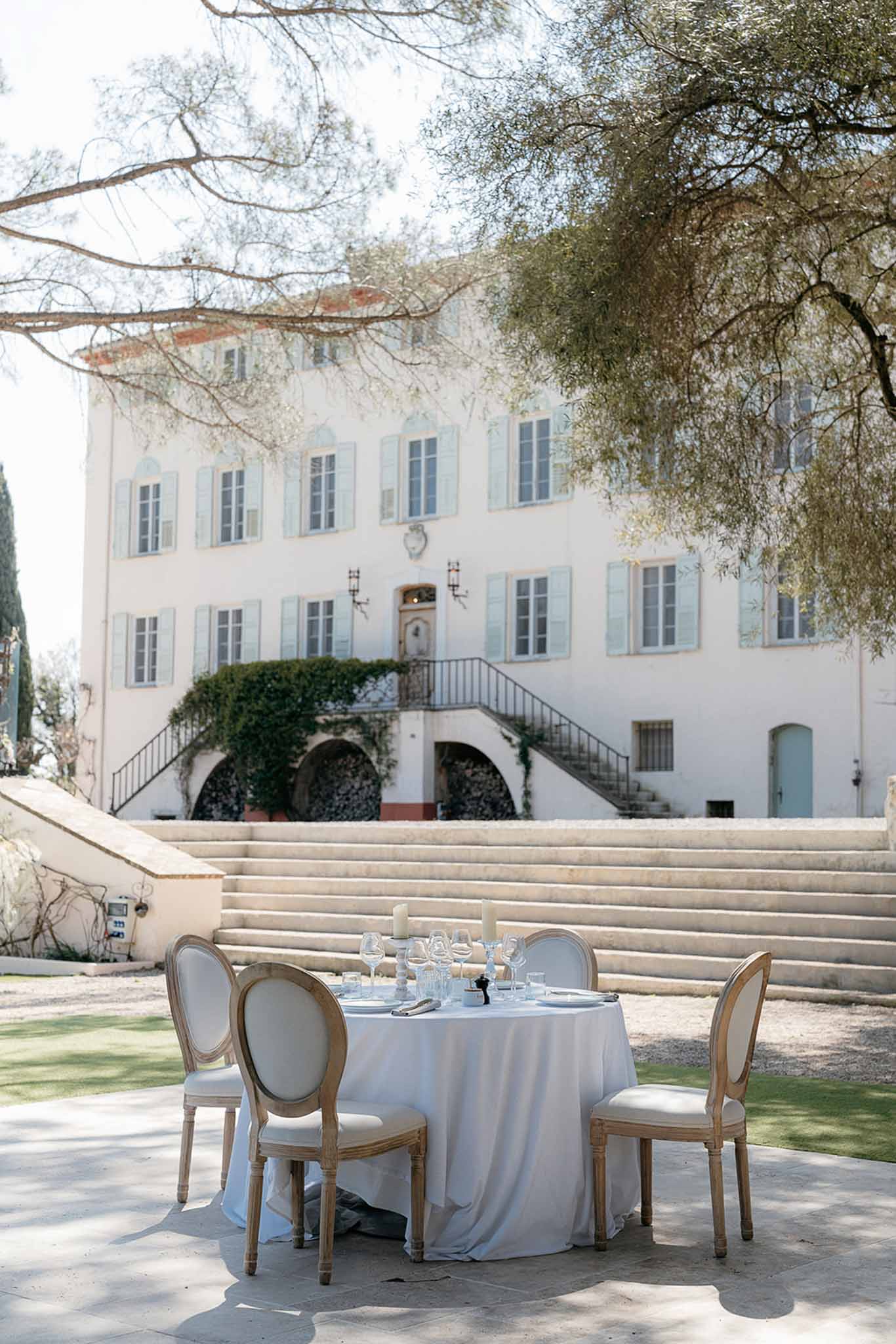 Sweetheart table with white linen and candles set before a Provencal manor house with blue-grey shutters and staircase