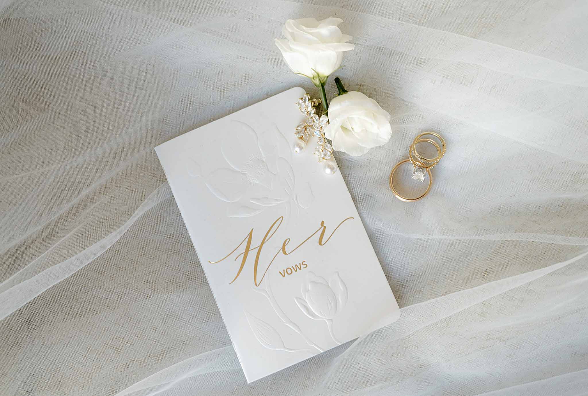 Flat lay of Her Vows booklet crystal pearl earrings gold rings and white roses on tulle veil