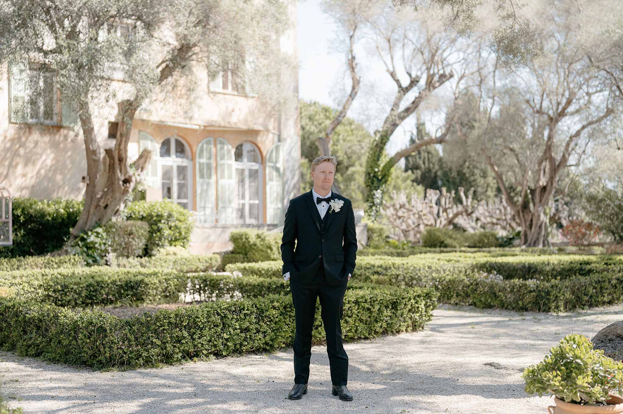 Groom in black tuxedo standing on gravel path in formal parterre garden with olive trees and ochre villa