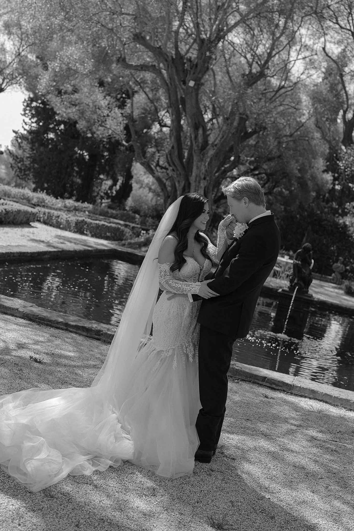 Black and white groom kissing bride hand beside reflecting pool with lace gown train spread on ground