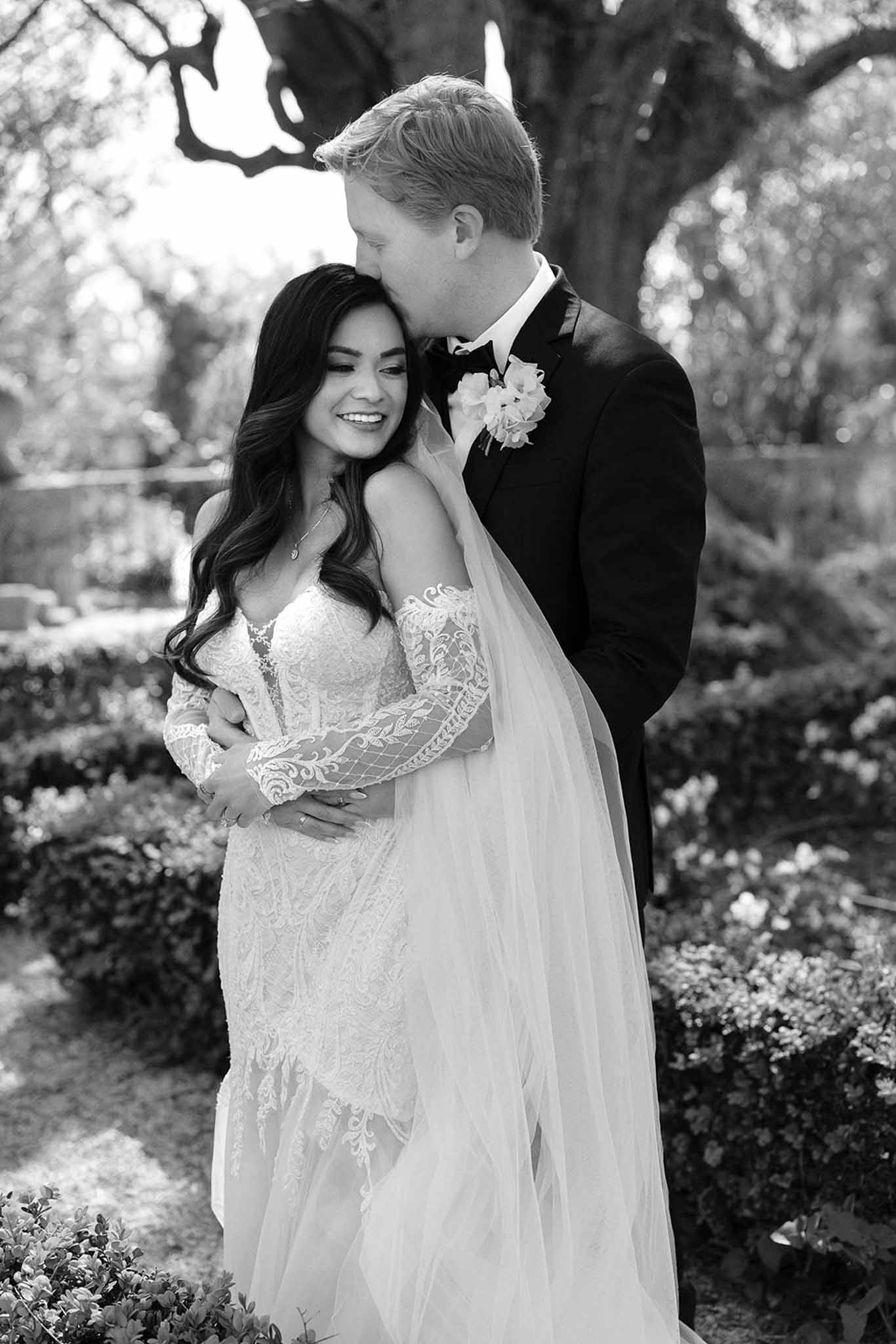 Black and white portrait of groom kissing bride's temple in lace gown with veil in formal garden