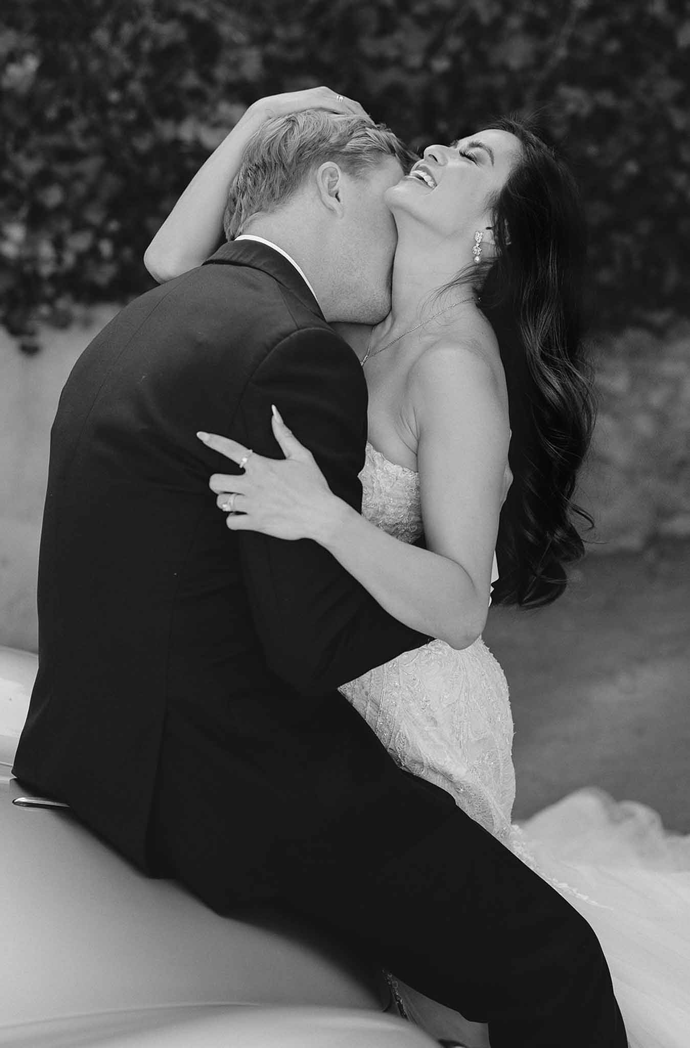 Groom kissing bride's neck as she laughs in strapless lace gown with crystal earrings in B&W
