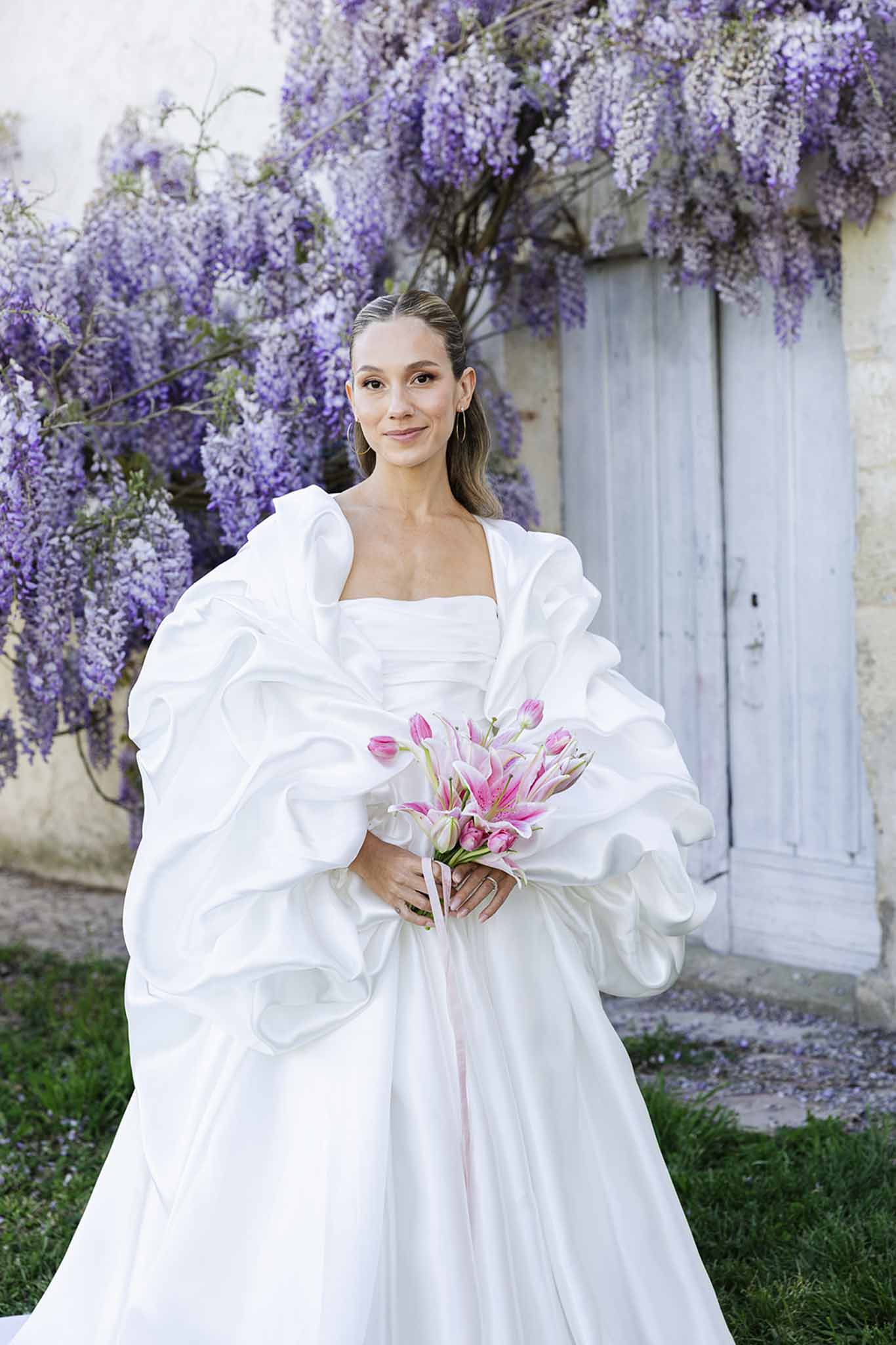 Bride in white dress holding pink lily bouquet in garden courtyard with wisteria