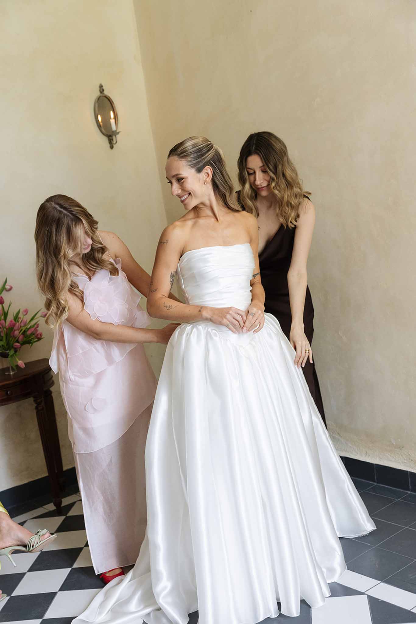 Bride in ivory gown being helped by attendants in elegant dressing room with checkered floors