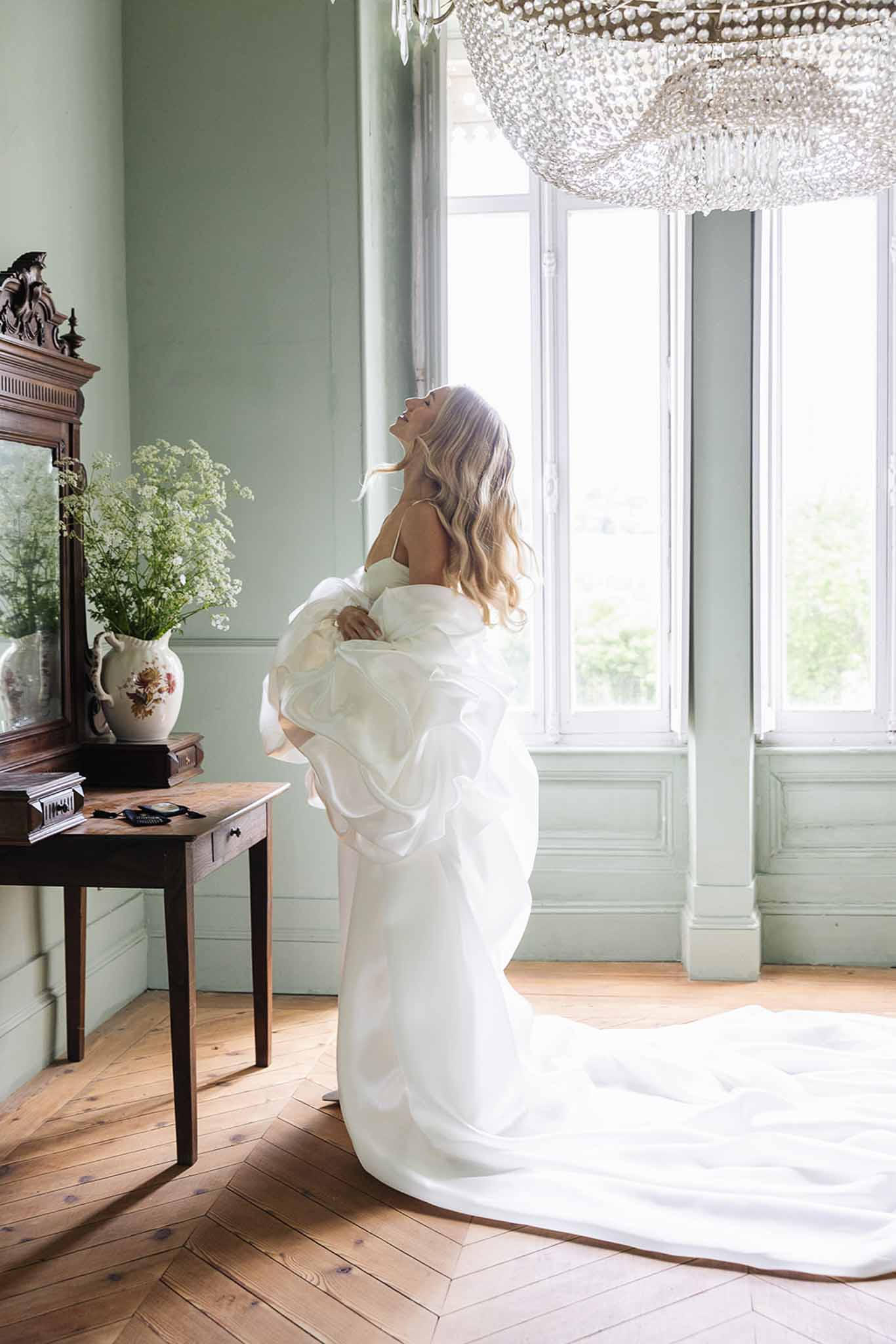 Bride in ivory wedding dress standing in elegant sage-green bridal suite with crystal chandelier