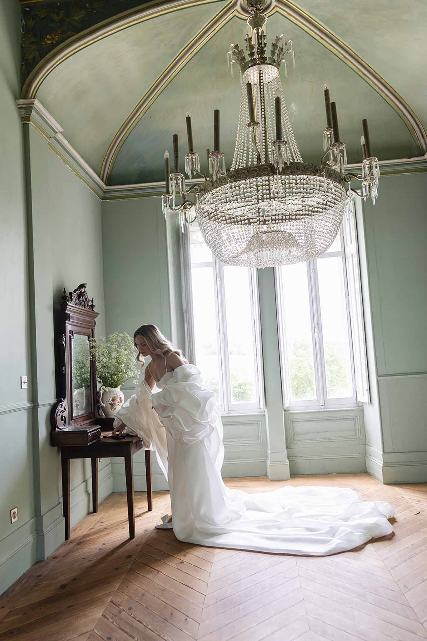 Bride in white off-shoulder ruffle-sleeve gown with cathedral train in sage green panelled chateau room with chandelier