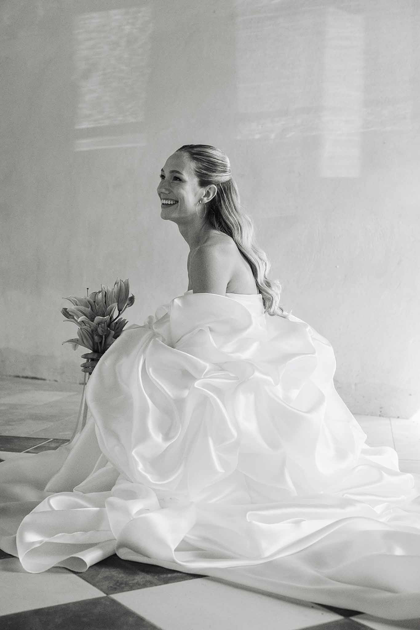 Bride in sculptural wedding dress seated against white wall holding bouquet