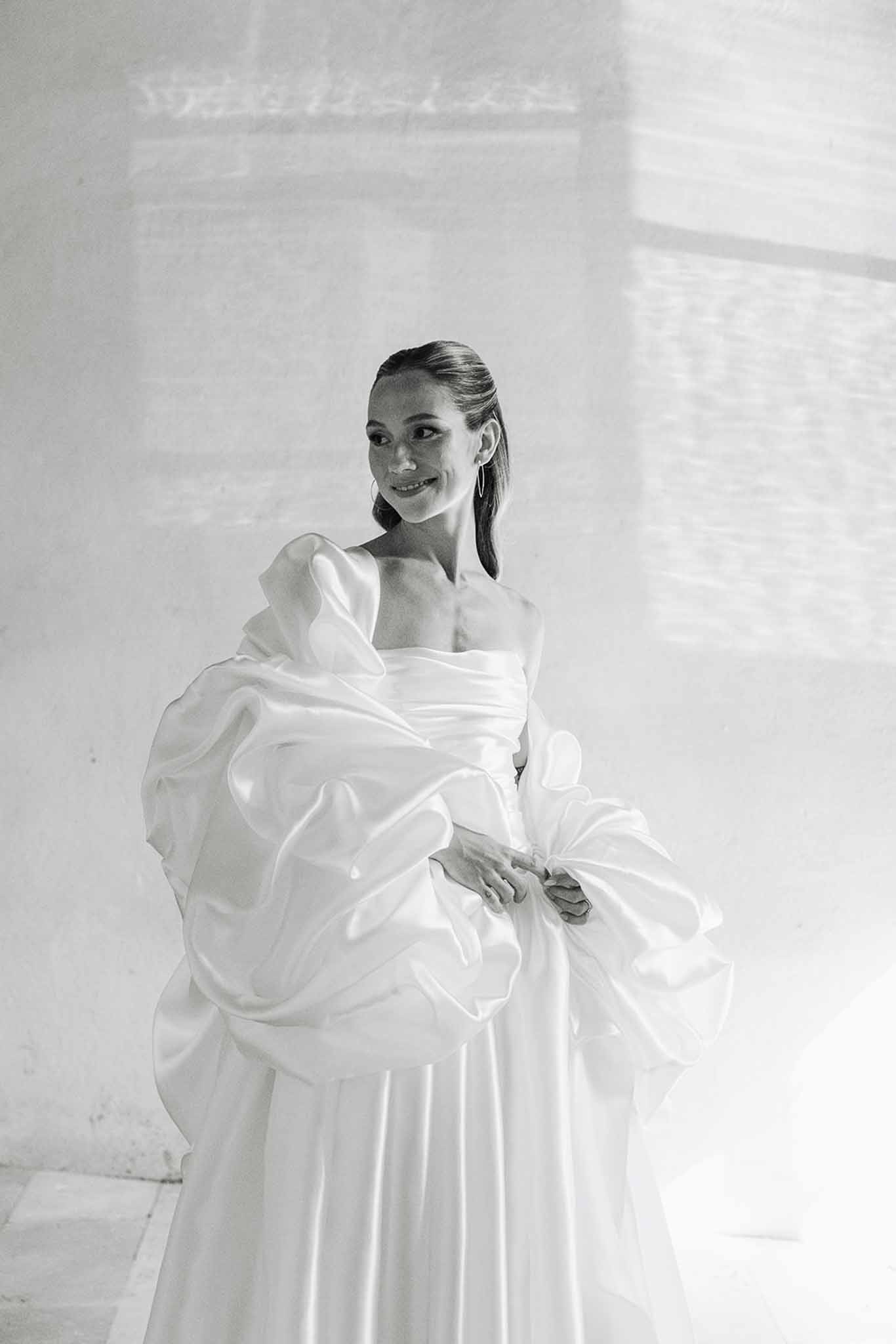 Bride in off-shoulder wedding dress with dramatic sleeves posing against white wall in black and white portrait
