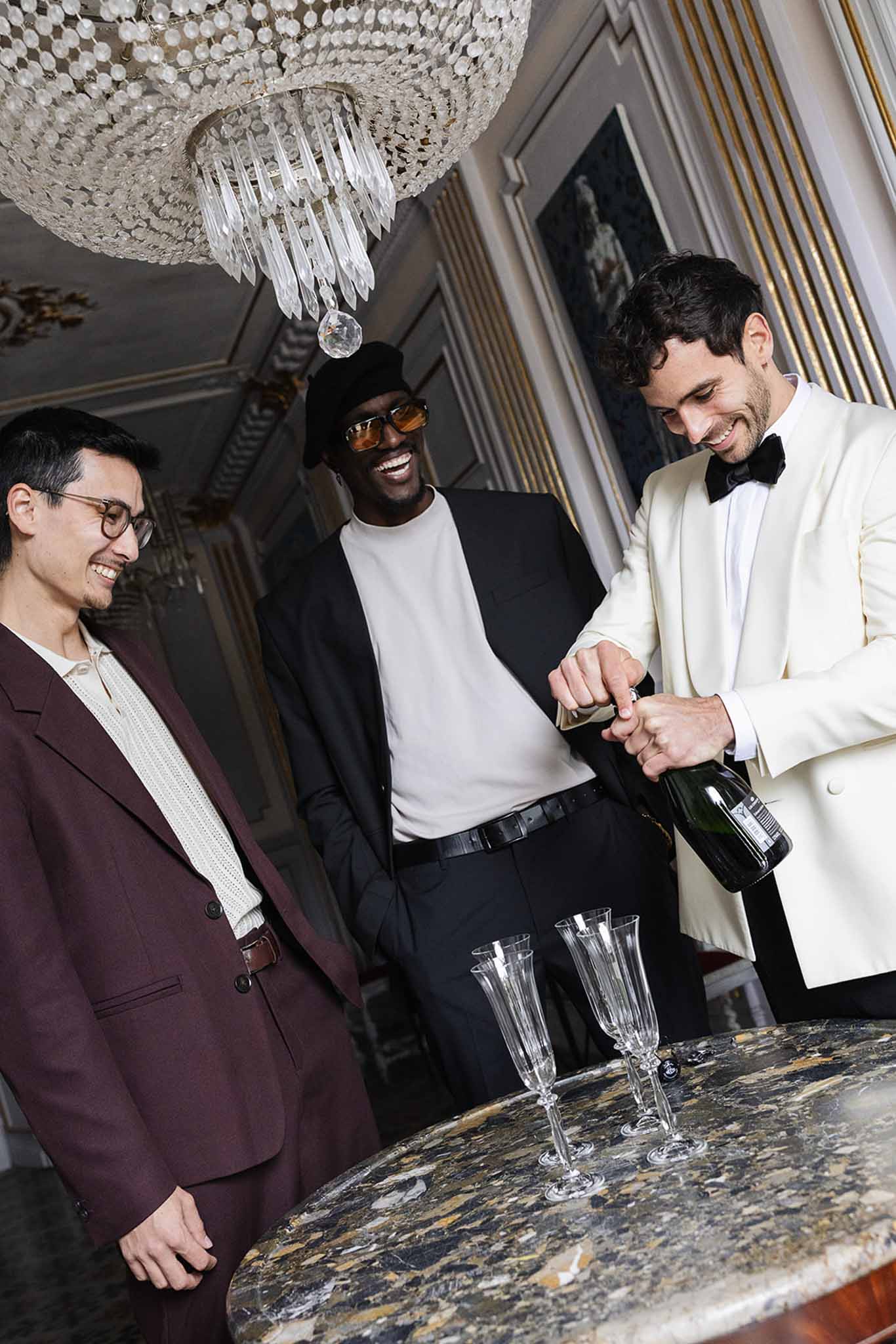 Groom opening champagne bottle with male guests laughing at indoor reception bar with crystal chandelier