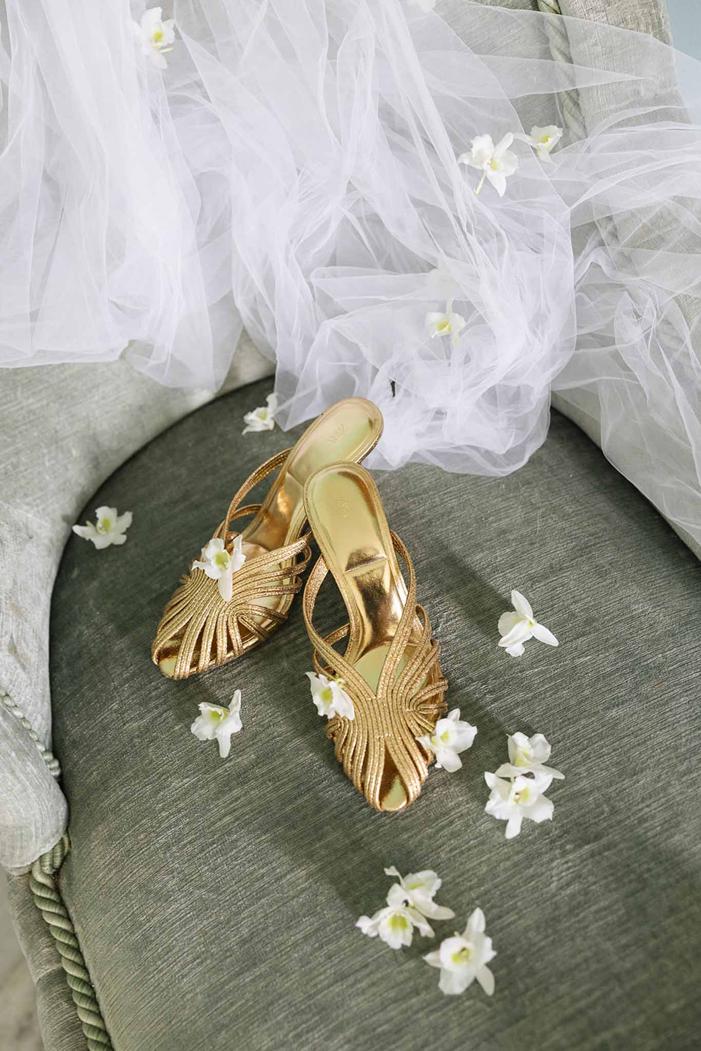 Bridal accessories flat lay with gold heeled sandals and white flowers on sage green linen