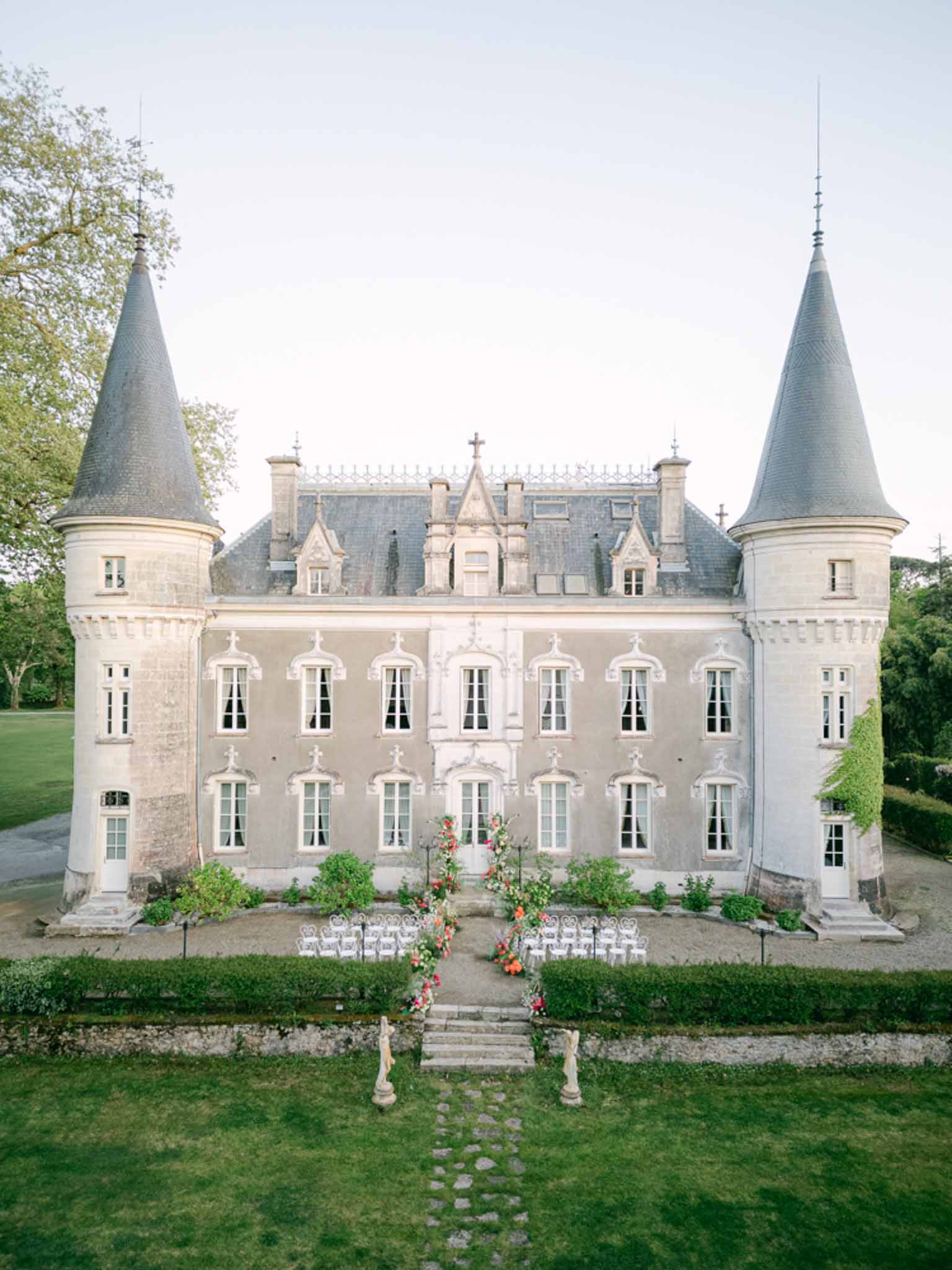 Aerial view of turreted chateau with white chair ceremony setup and coral floral arch on forecourt