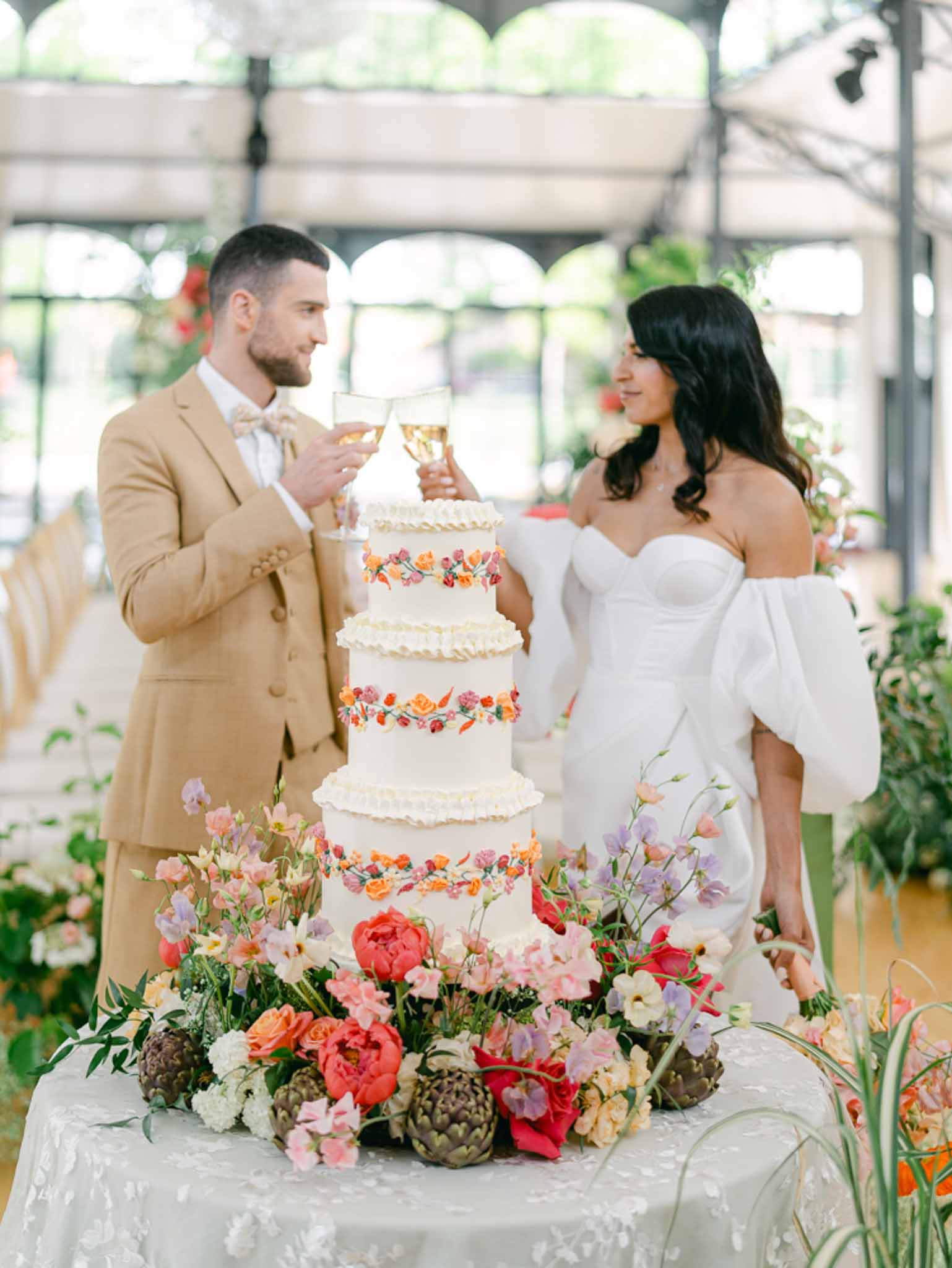 Couple toasting beside five-tier ivory cake with coral floral motifs and garden flower arrangement in greenhouse