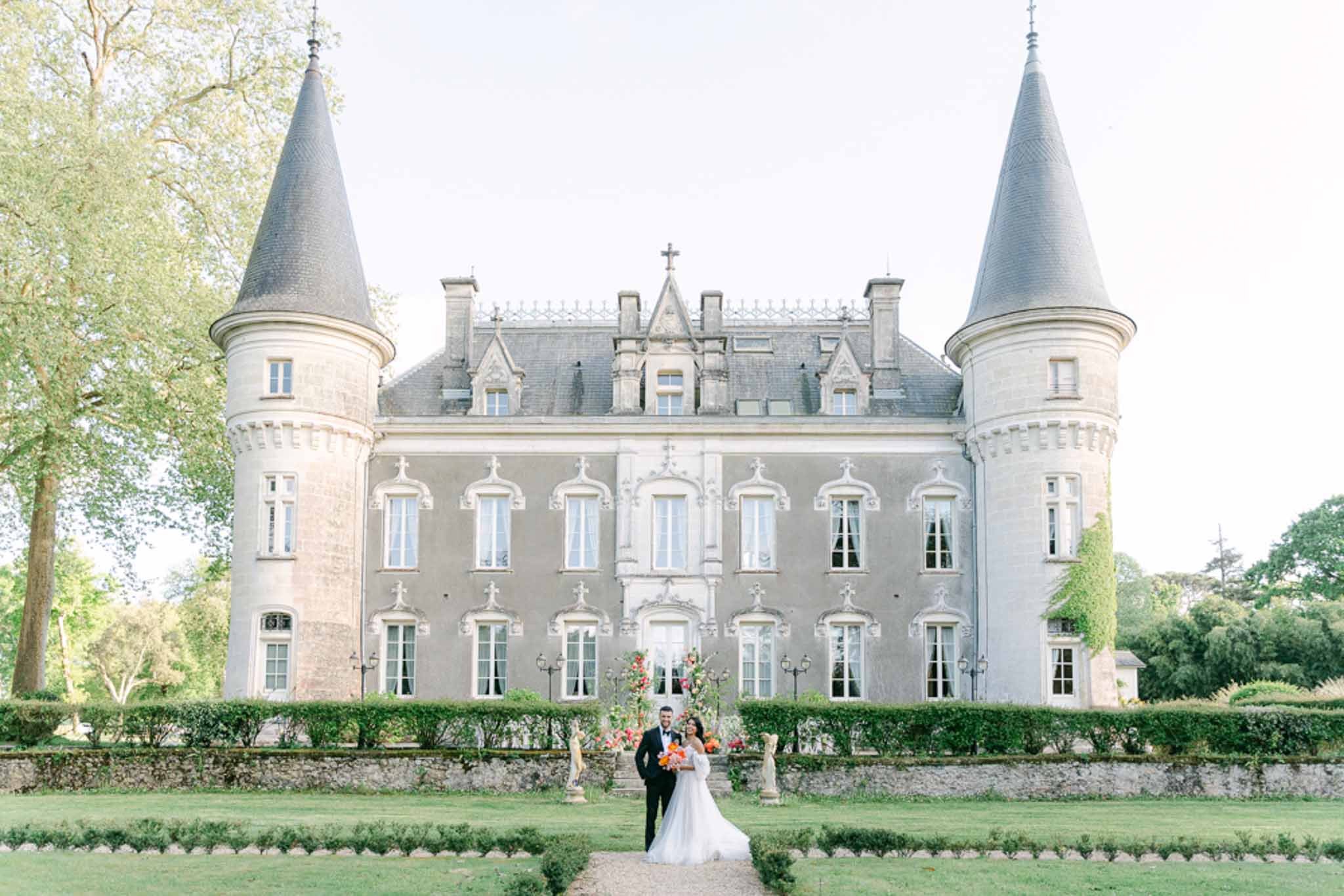 Bride and groom pose before a turreted chateau with an orange and red floral installation at the entrance