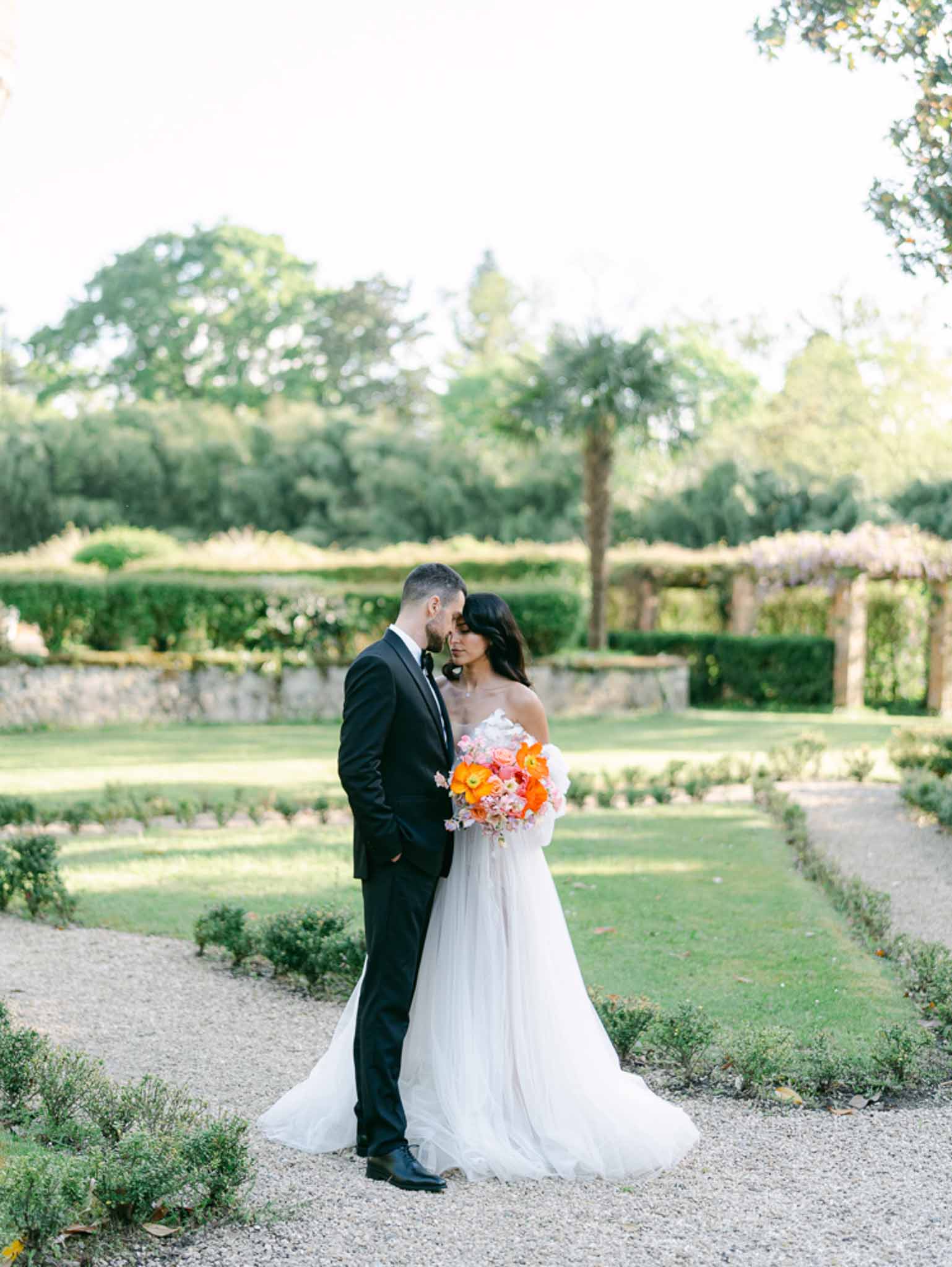 Couple foreheads touching in formal garden bride in tulle ballgown with orange poppy and coral bouquet groom in navy tuxedo