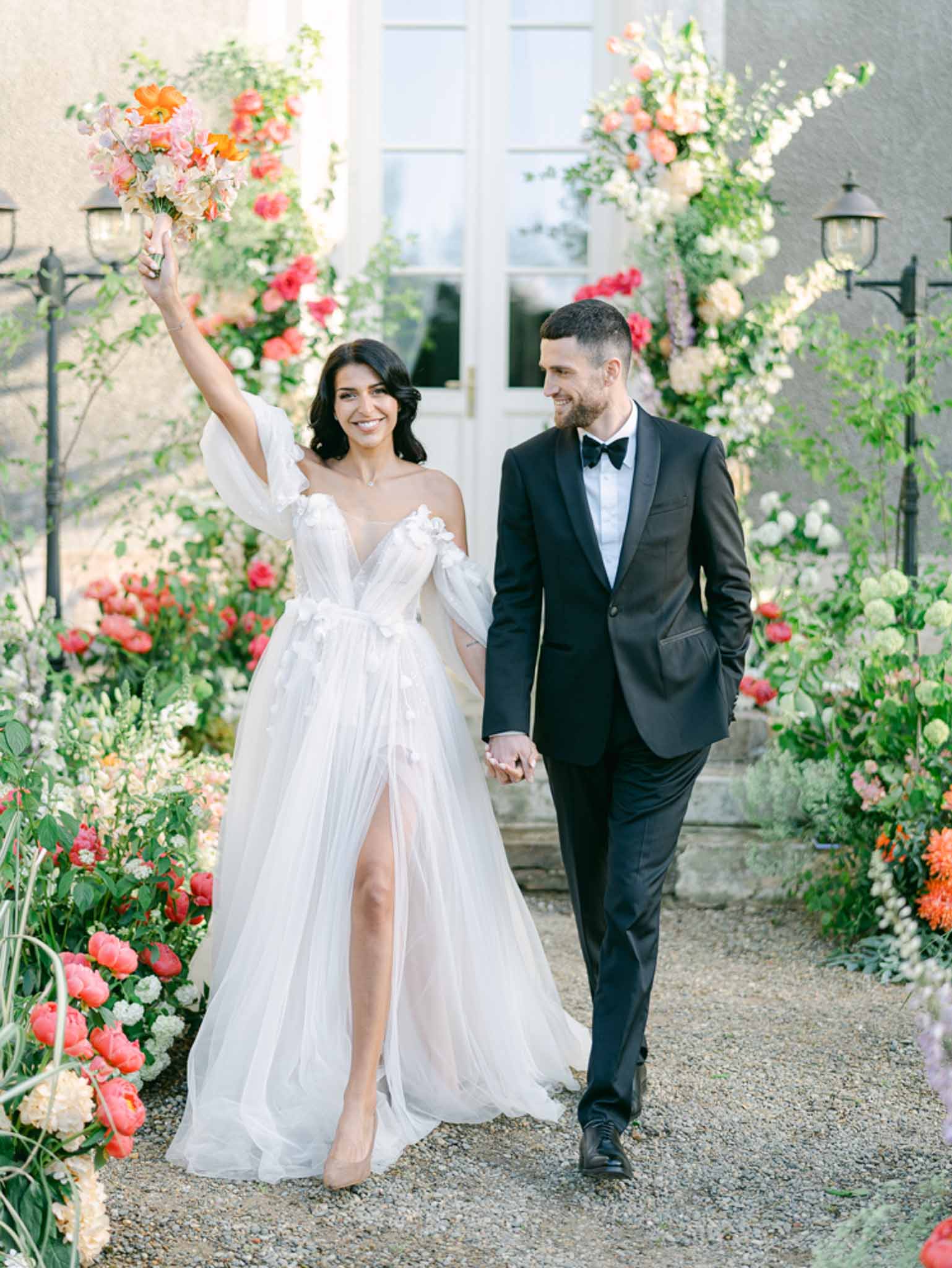 Couple walking hand-in-hand along gravel path flanked by coral and orange floral installations