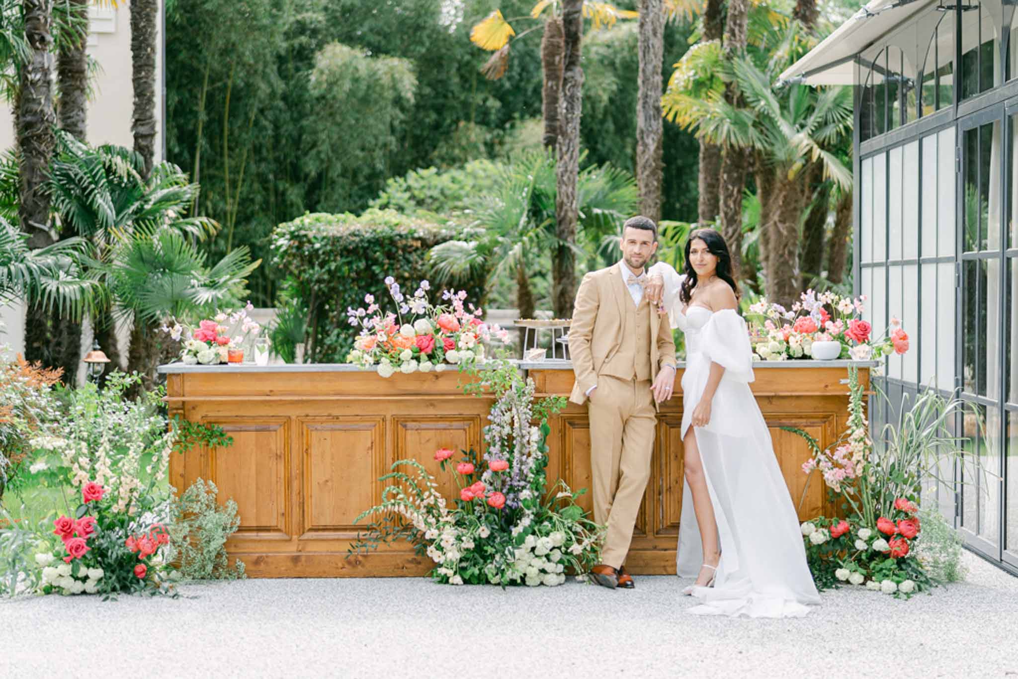 Bride and groom posing in front of outdoor bar decorated with coral roses, pink blooms, and trailing greenery