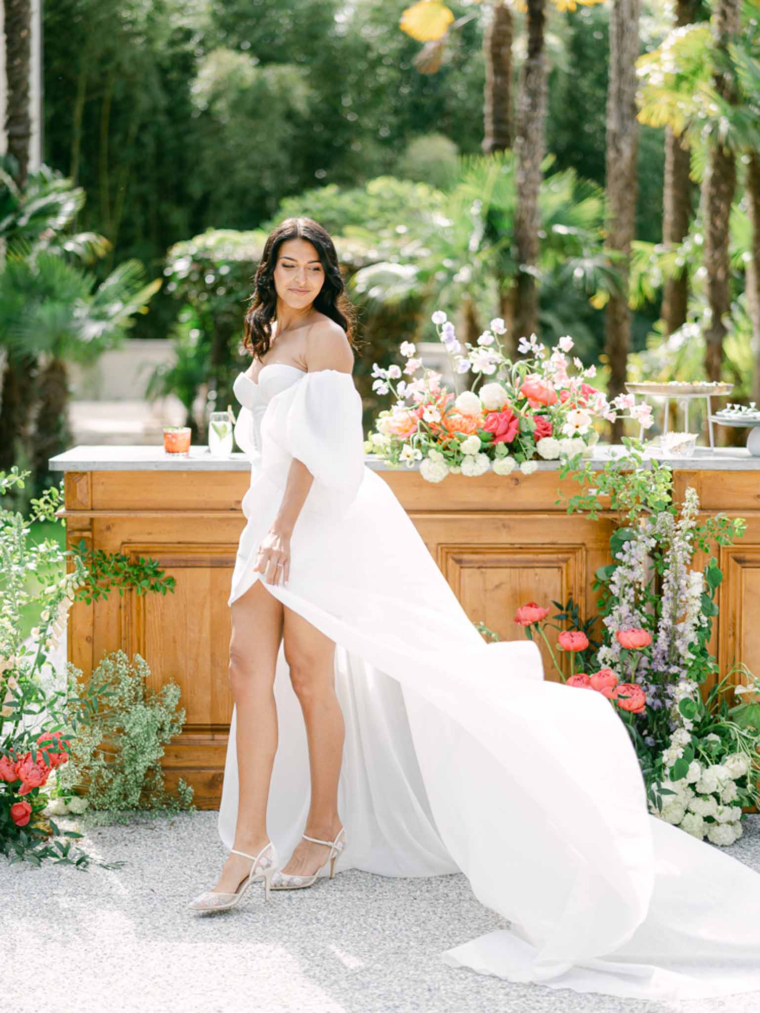 Bride in off-shoulder gown with puff sleeves beside floral bar with coral peonies and lavender delphinium