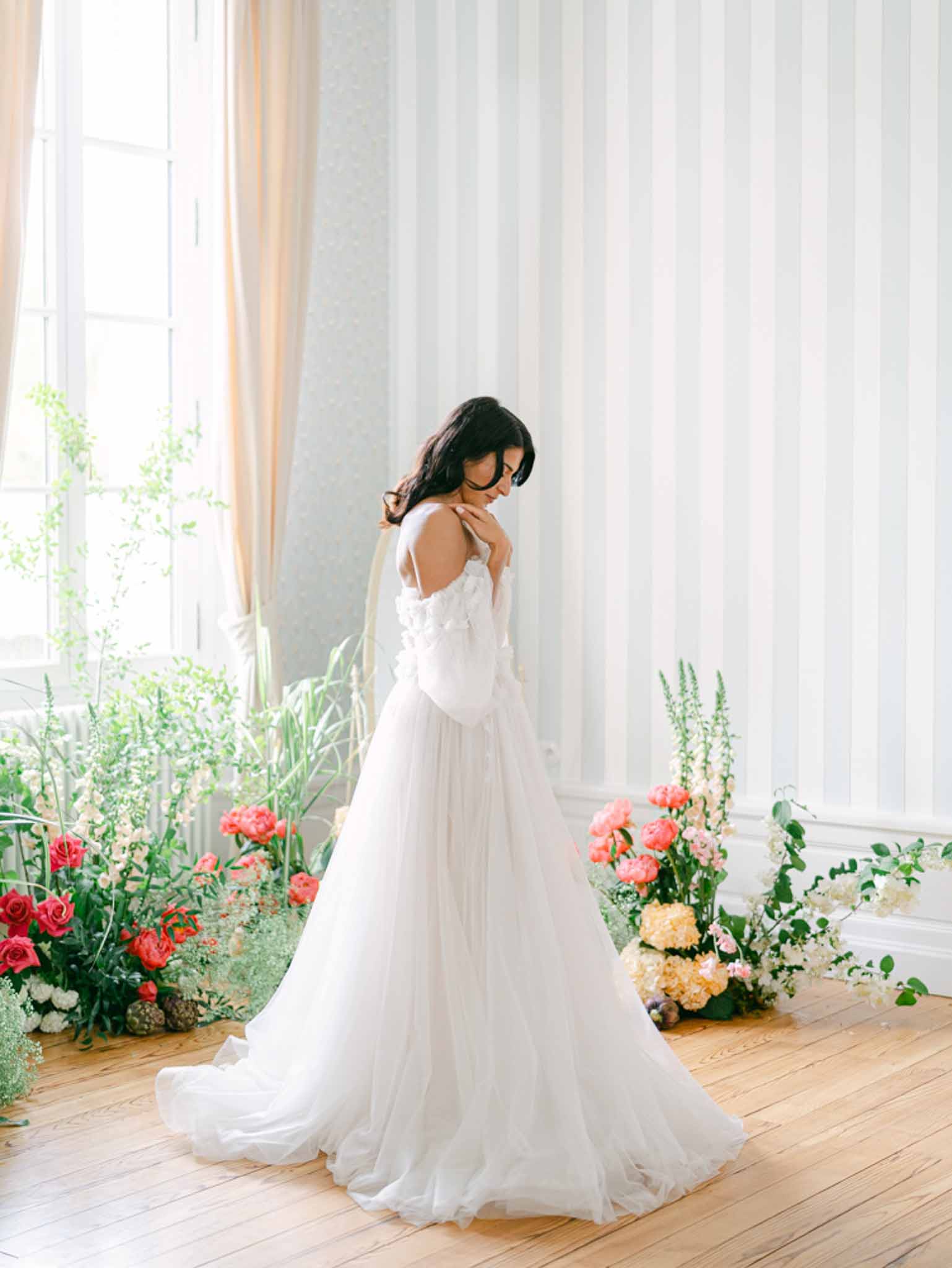 Bride in off-shoulder floral applique gown surrounded by coral red and white ground floral arrangements in bright panelled...