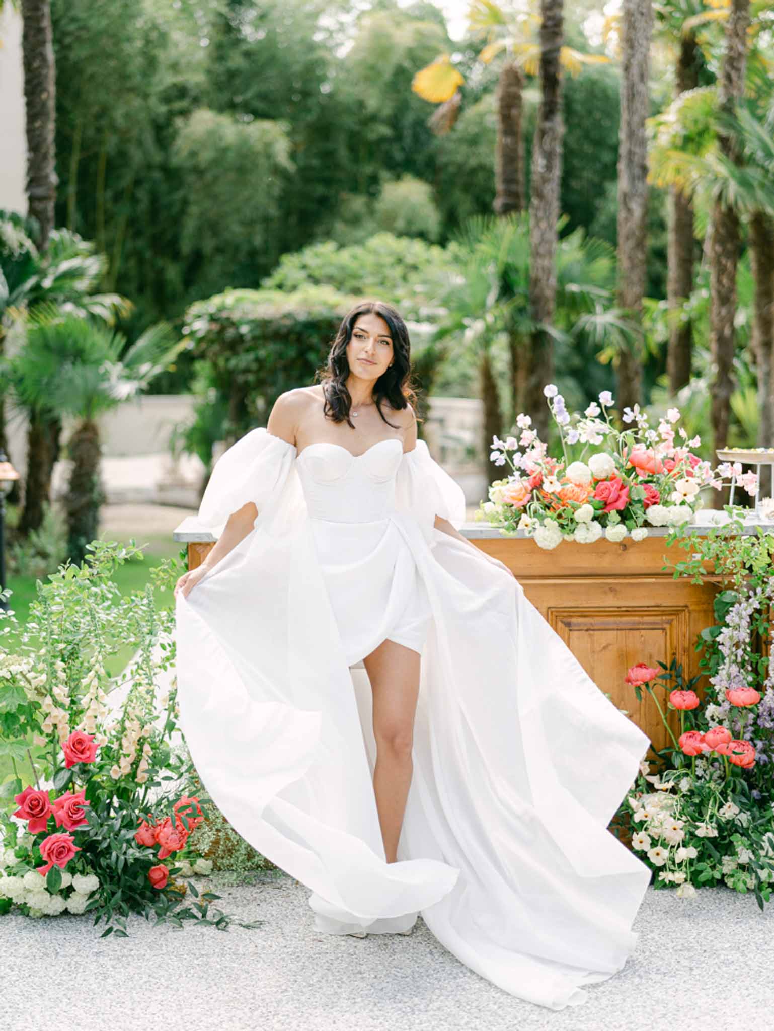 Bride in puff-sleeve gown holds skirt wide before lush coral rose and lavender bar arrangement with palms