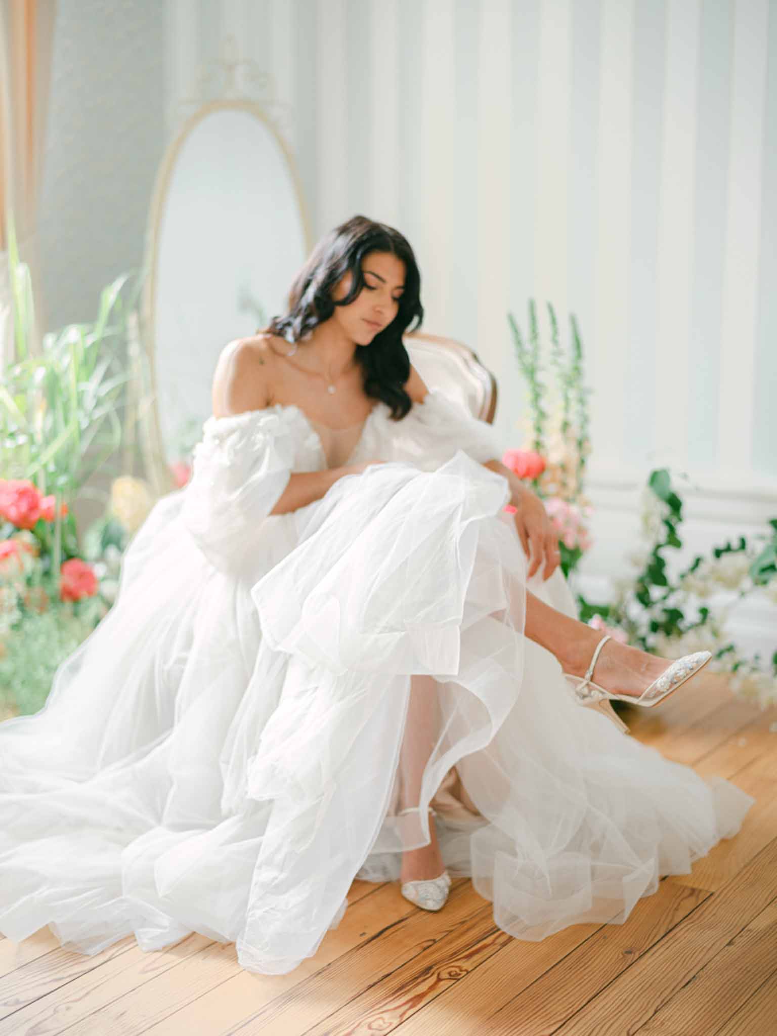 Bride seated on a gilded chair wearing a ruffled organza ballgown surrounded by coral peonies and pink foxglove