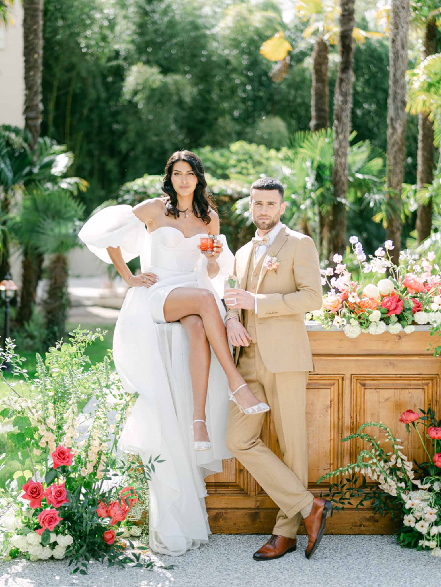 Bride seated on rustic wooden bar and groom standing beside her holding drinks at floral-decorated cocktail station